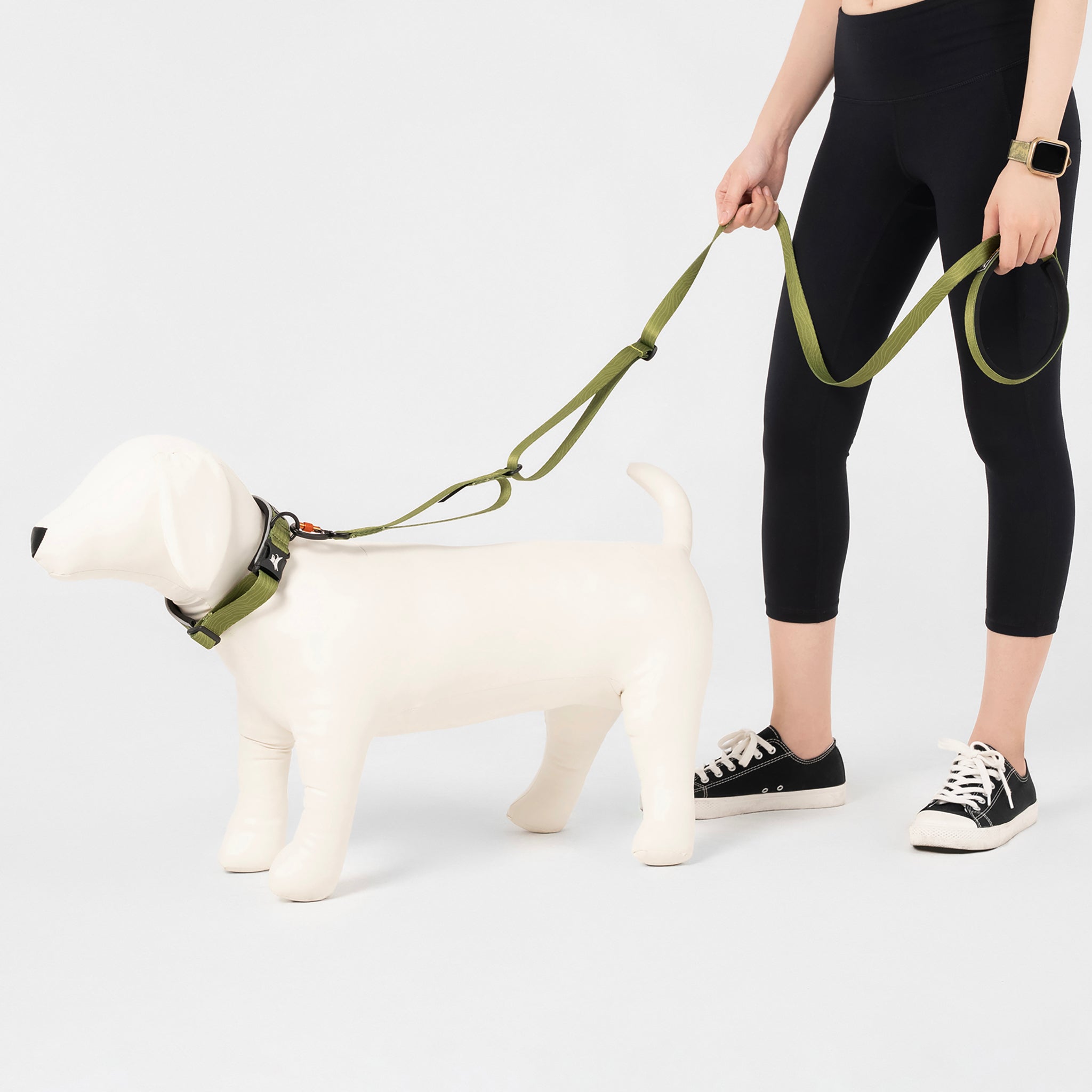 Person holding the OllyDog Flagstaff Adjustable Leash in Cactus Woodgrain, attached to a dog mannequin. The leash features an adjustable waist belt, padded handle, sturdy polyester webbing, and swivel carabiner, designed for hands-free and secure dog walking #color_cactus woodgrain