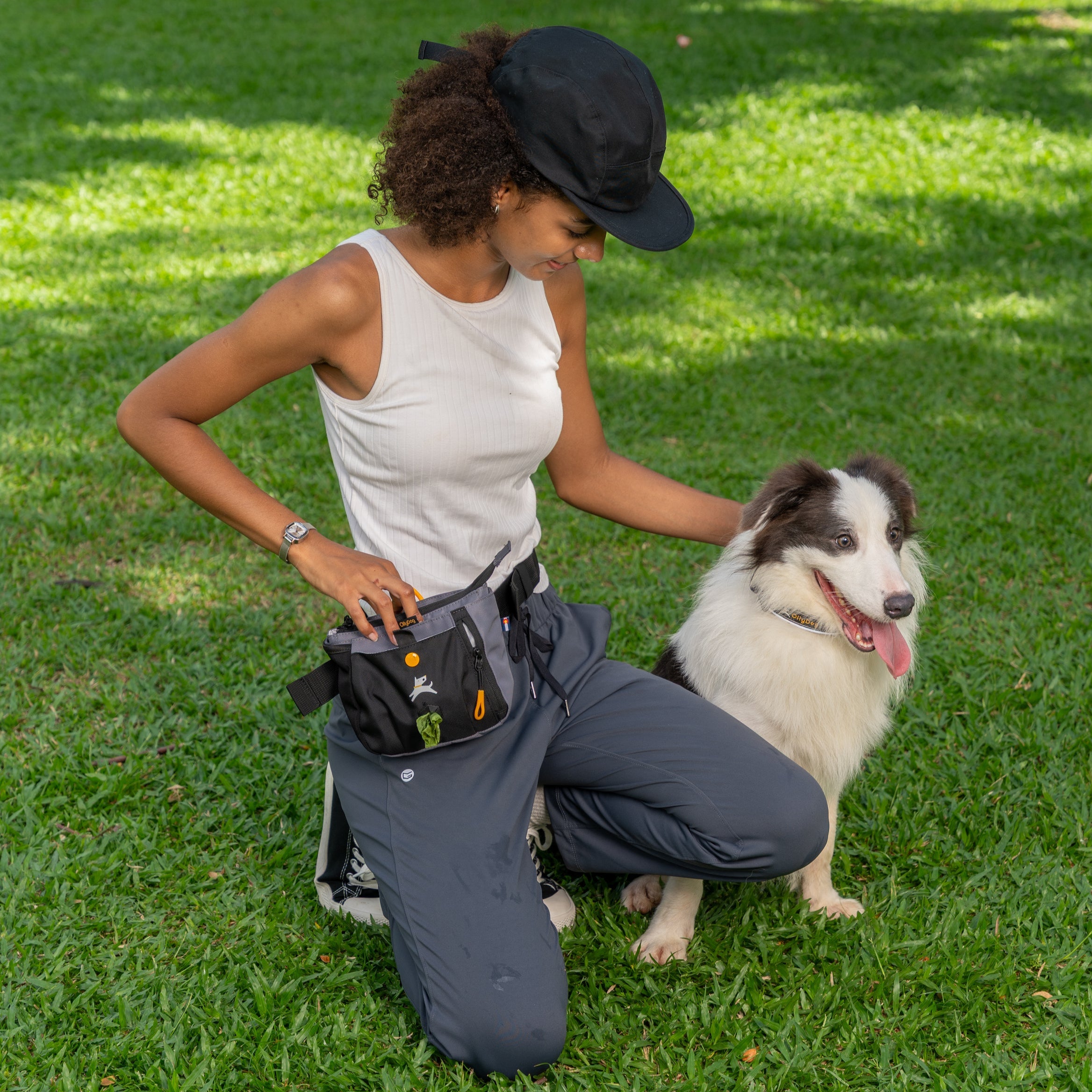 Woman kneeling on grass during dog training session, wearing the OllyDog Backcountry Day Bag in Castle Rock with waste bags visible #color_castle rock