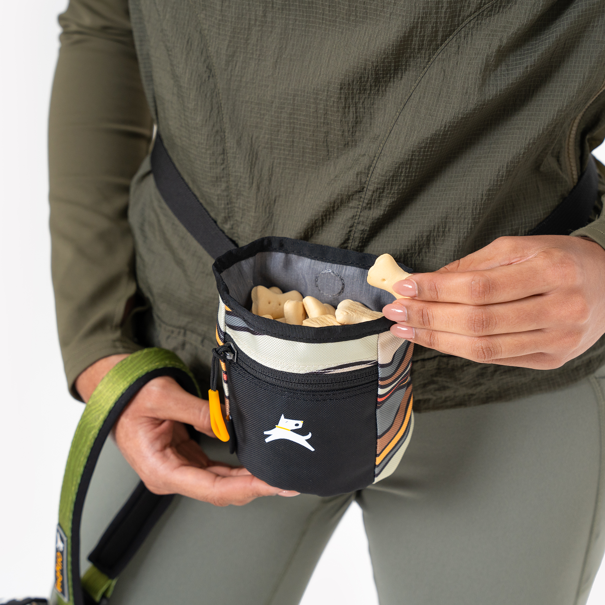 A person wearing a striped OllyDog Goodie Treat Bag around their waist, with their hands reaching inside to grab a light-colored dog treat, demonstrating easy access during training #color_ventura '73