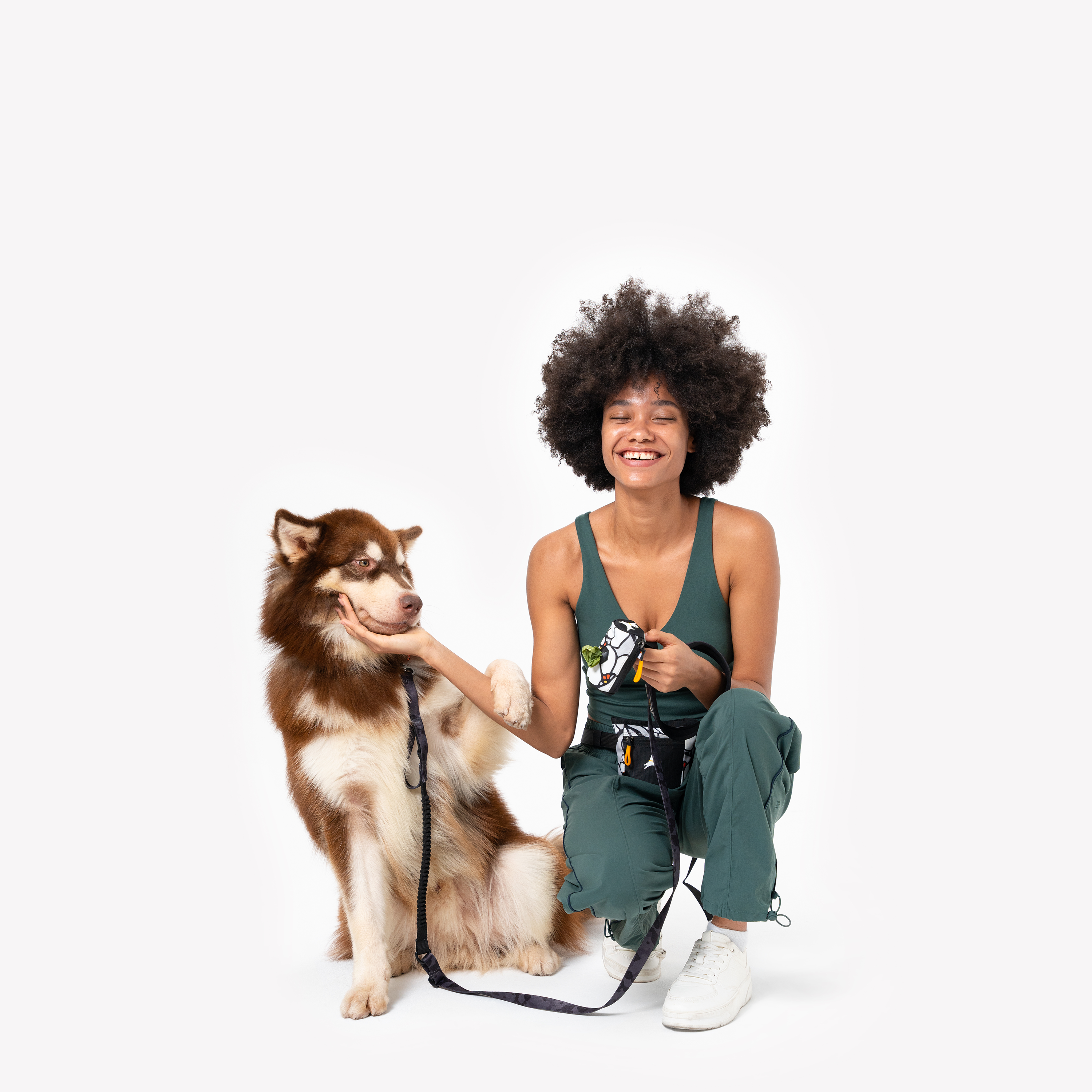 Smiling woman kneeling beside a brown and white dog, holding an OllyDog Goodie Treat Pouch in Magnolia color, wearing it on her waist #color_magnolia