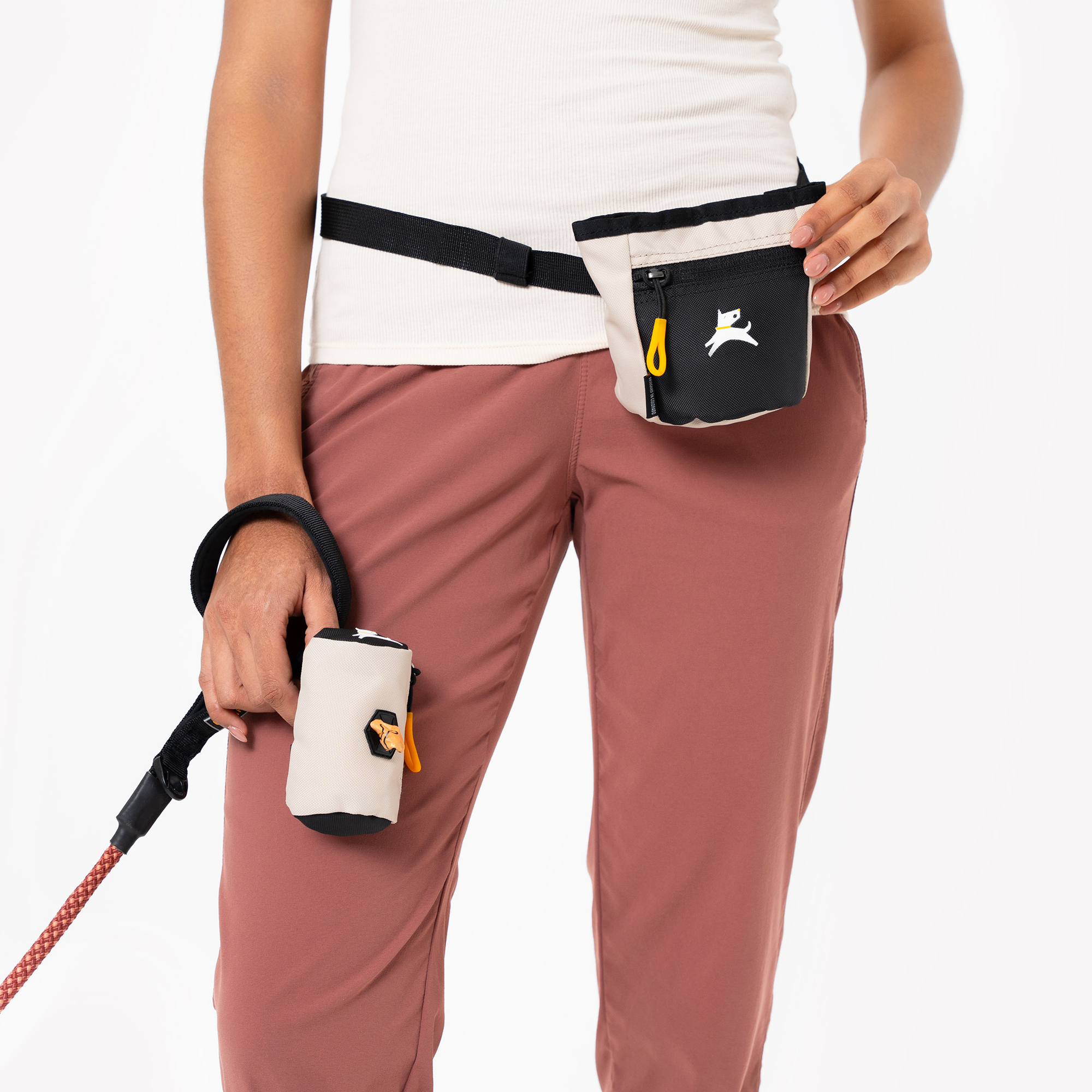 Person wearing a treat pouch clipped to a waist belt and holding a matching waste bag holder while walking a dog. Both accessories are in a black and cream color combination with yellow zipper accents #color_con leche
