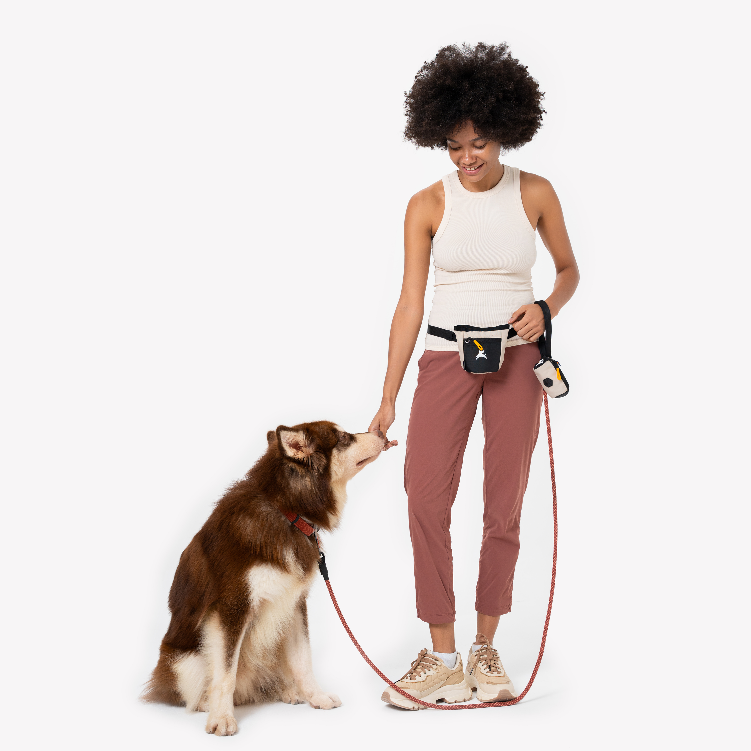 Smiling woman walking her dog, wearing a treat pouch on her waist and holding a matching waste bag holder. The dog sits attentively on a leash, looking up at her #color_con leche
