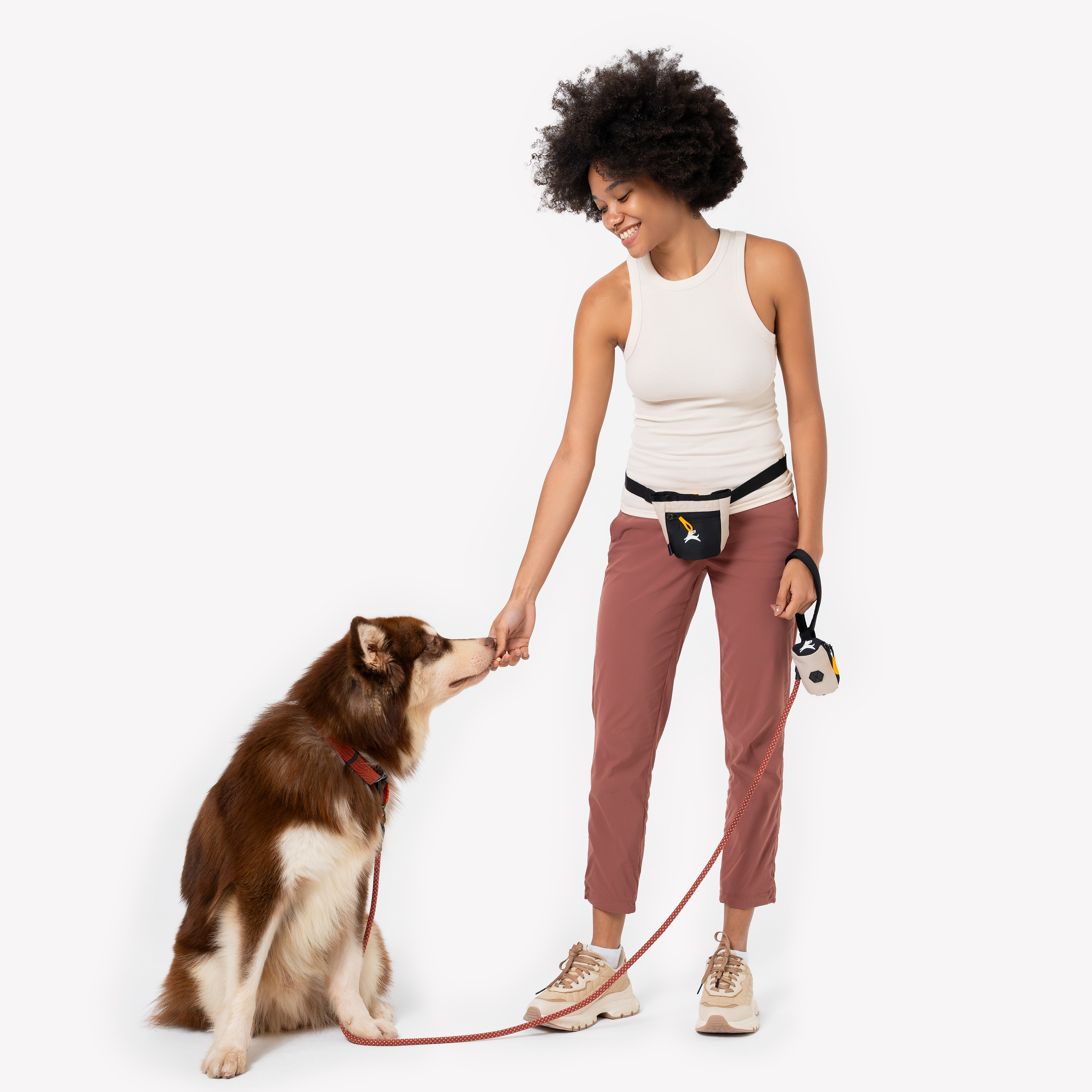 Woman smiling and giving a treat to her sitting dog during a walk. She wears a treat pouch at her waist and holds a matching waste bag holder, both in a black and beige design #color_con leche