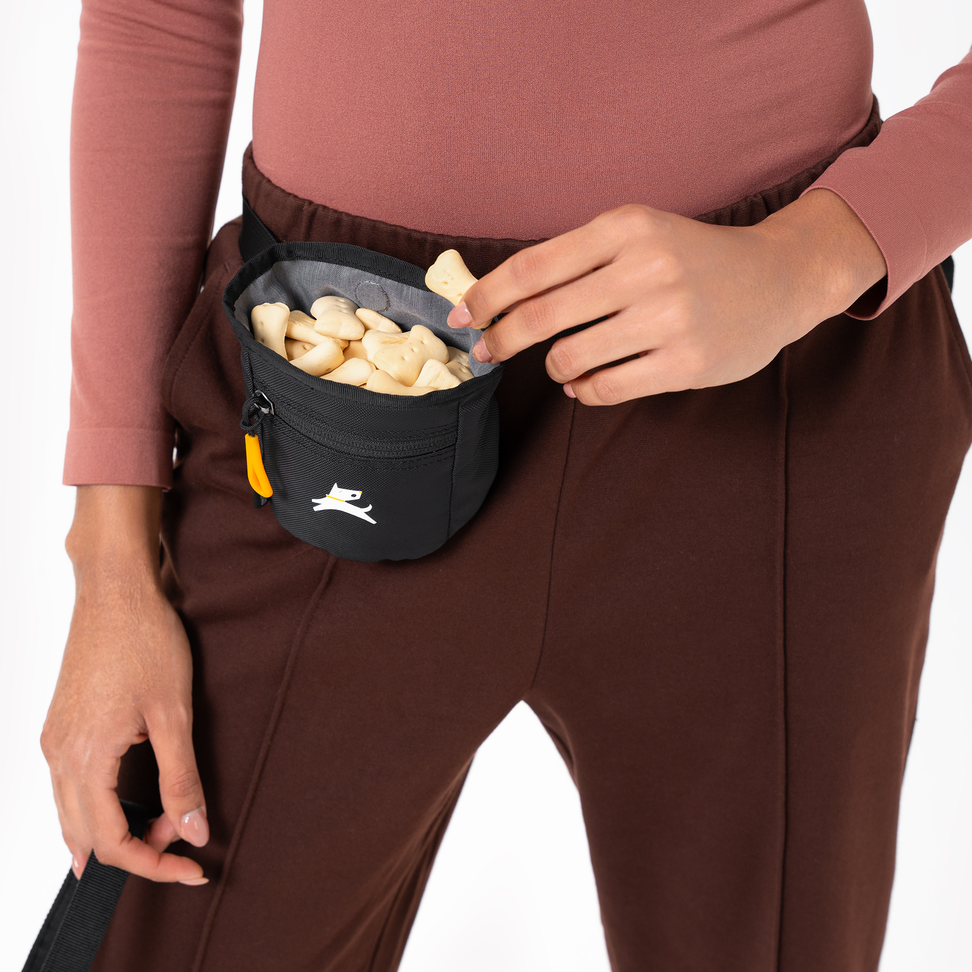 Close-up of a person wearing a black dog treat pouch filled with bone-shaped treats, attached at the waist. One hand is reaching in to grab a treat, highlighting easy access during training #color_raven s'25