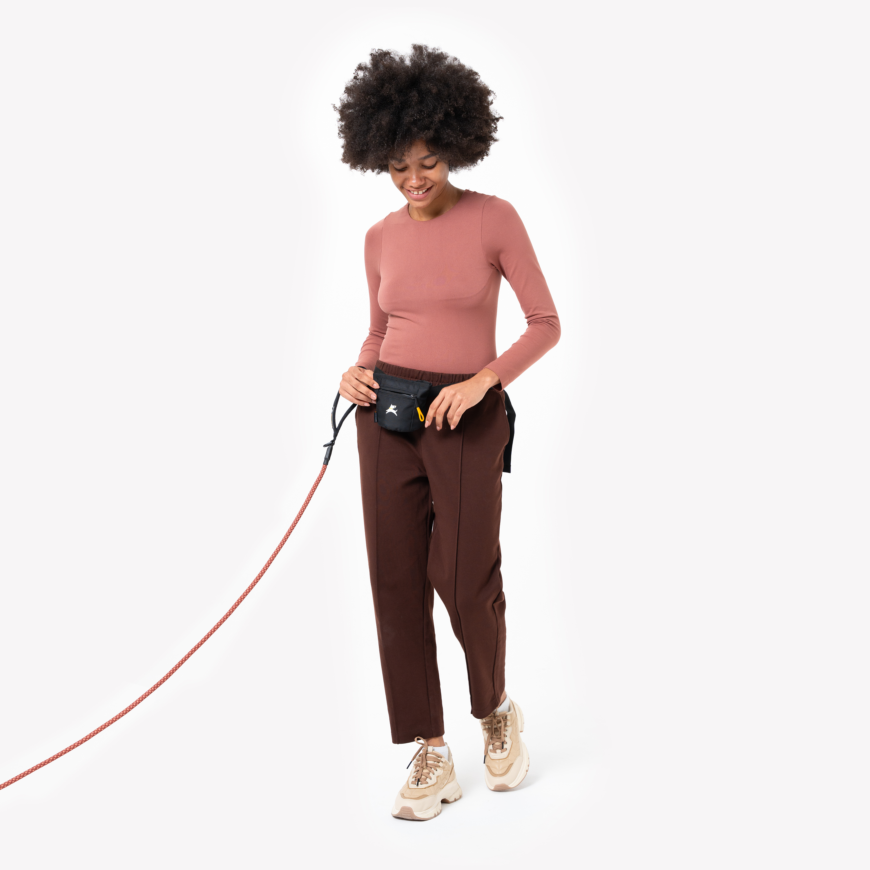 Woman walking a dog while wearing a black OllyDog Goodie Treat Pouch in Raven color on her waist. She is smiling and holding the leash with one hand while reaching into the pouch with the other #color_raven s'25