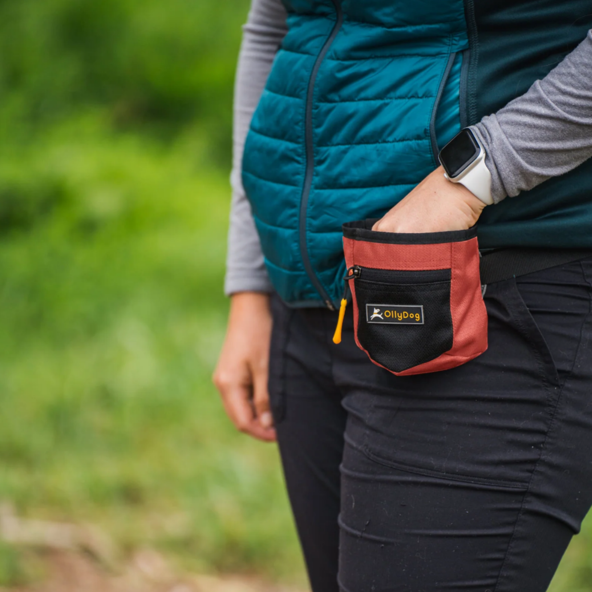 The image shows a person wearing a teal-colored puffy jacket and holding an OllyDog Goodie Treat Bag, which is a small treat pouch in a reddish-orange "Reef" color, attached to their waist. The pouch is likely used to carry dog treats or other pet supplies. #color_reef