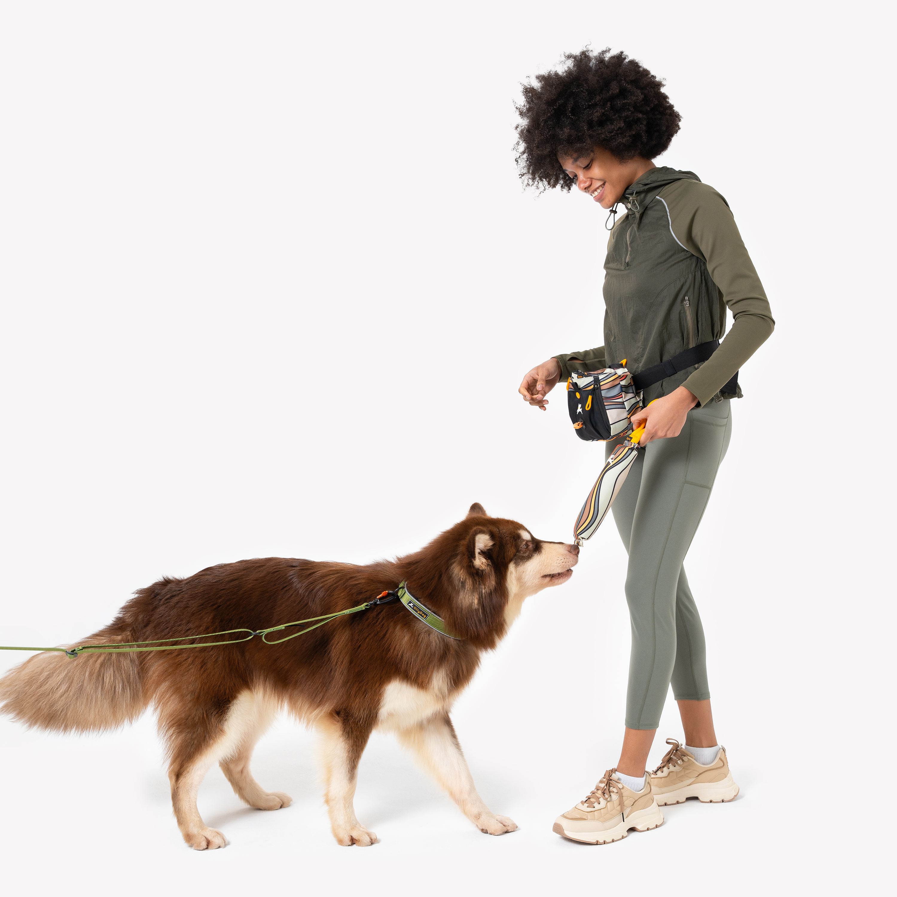 Model playing tug with a brown dog using the OllyDog Tugger interactive dog toy in Ventura '73 print, showing the retro wave pattern and sturdy handle, with the model wearing a green jacket and leggings #color_ventura '73
