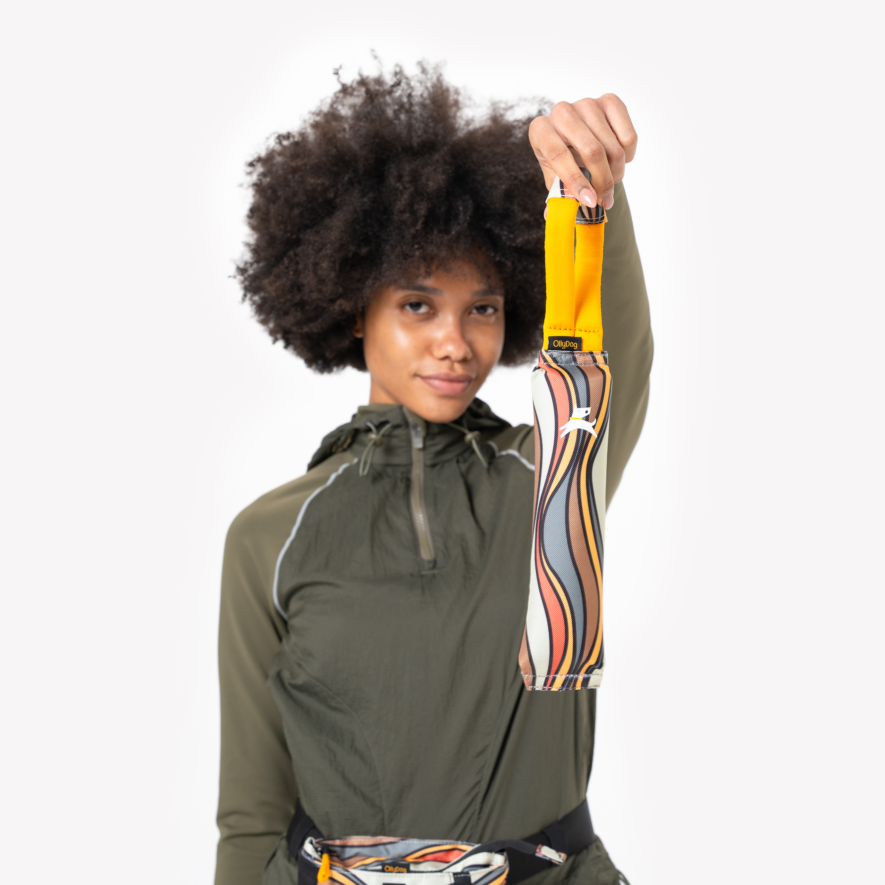 Model holding up the OllyDog Tugger interactive dog toy in Ventura '73 colorway, showcasing the retro swirl pattern, durable fabric, and sturdy yellow handle, with model wearing an olive green jacket #color_ventura '73