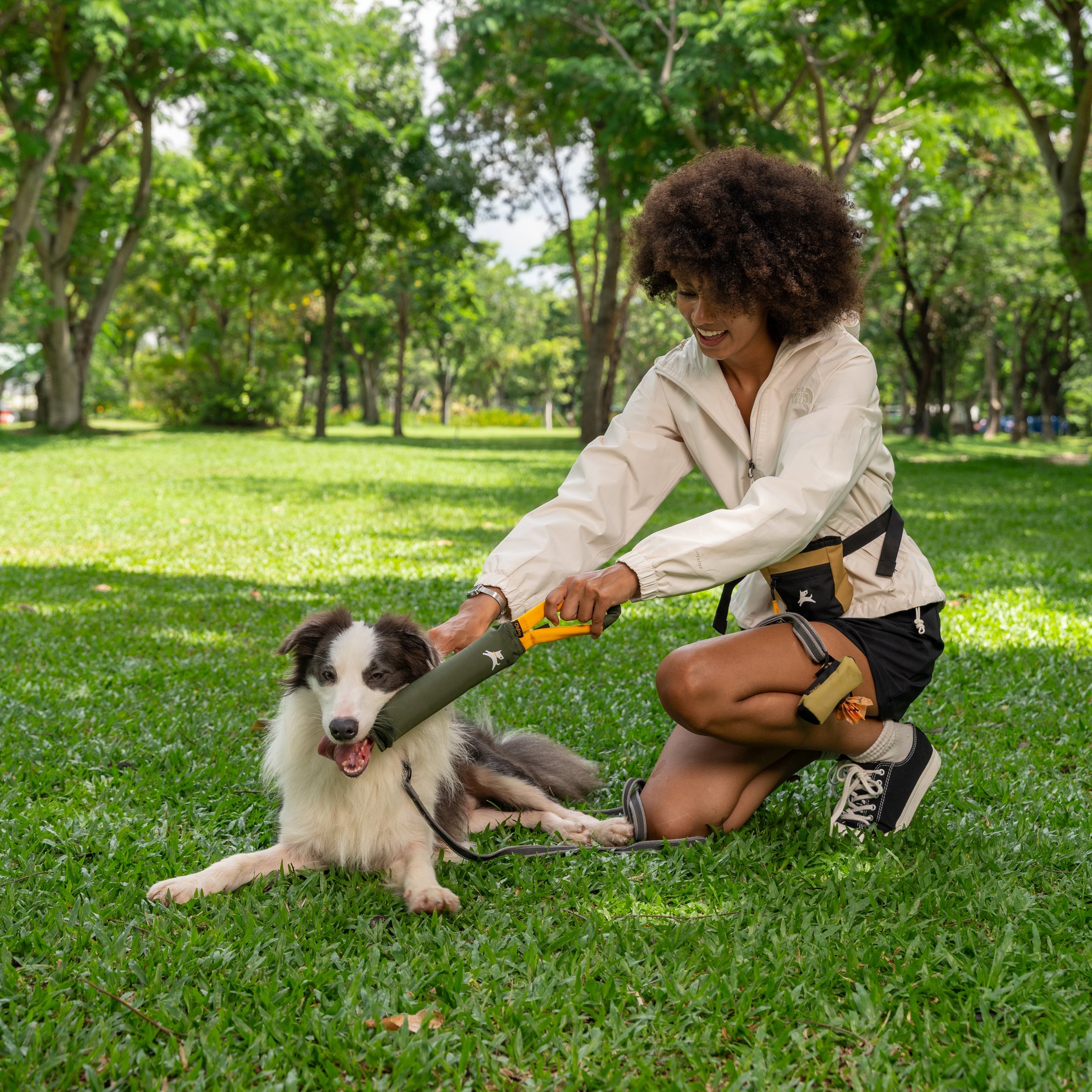 Woman playing with dog in the park using OllyDog Tugger toy, with the Scoop Pick Up Bag in Alchemy color clipped to the leash #color_alchemy