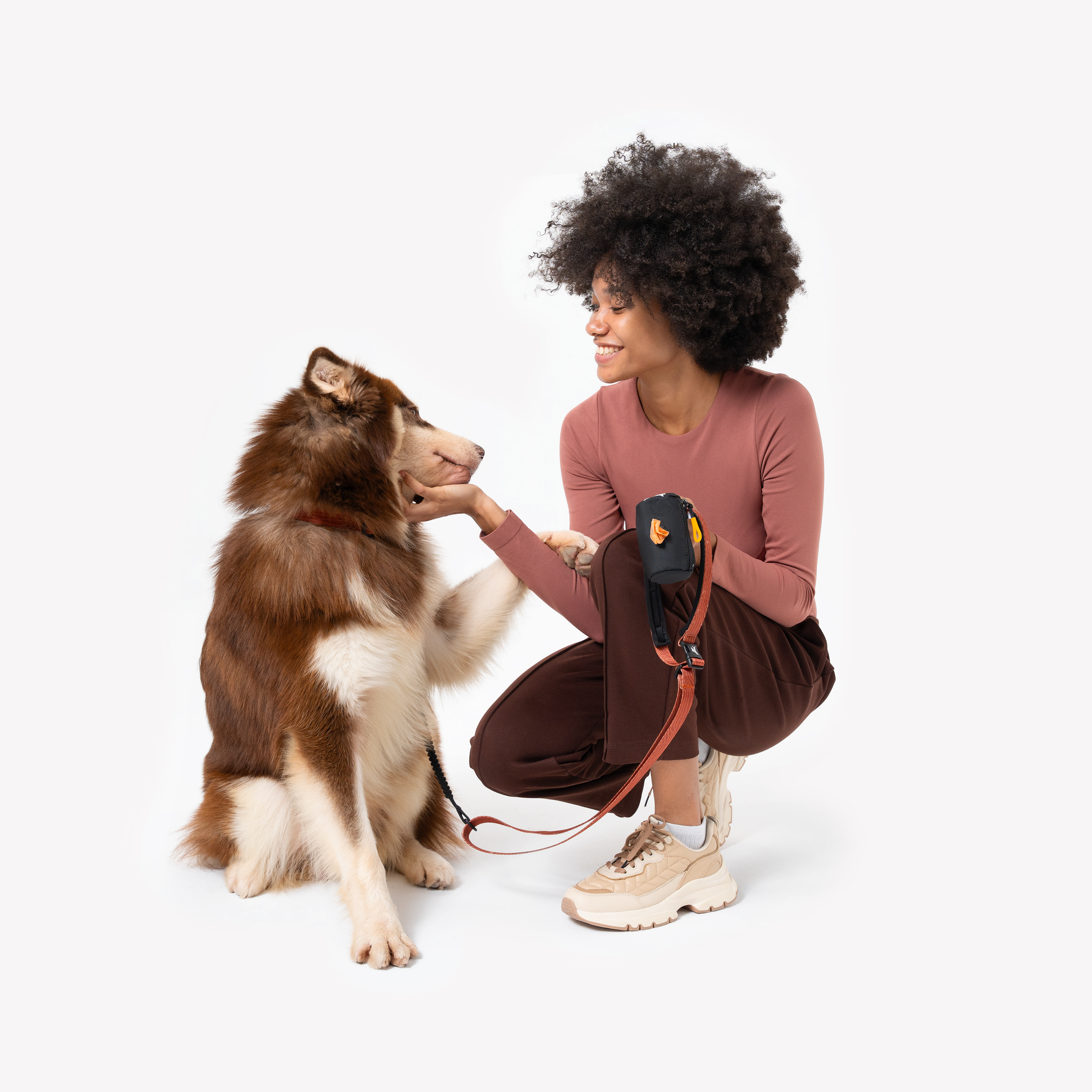 Woman crouching and smiling at a brown-and-white husky dog, holding an orange leash with a black waste-bag dispenser attached, wearing a pink long-sleeve top, brown pants, and beige sneakers #color_raven