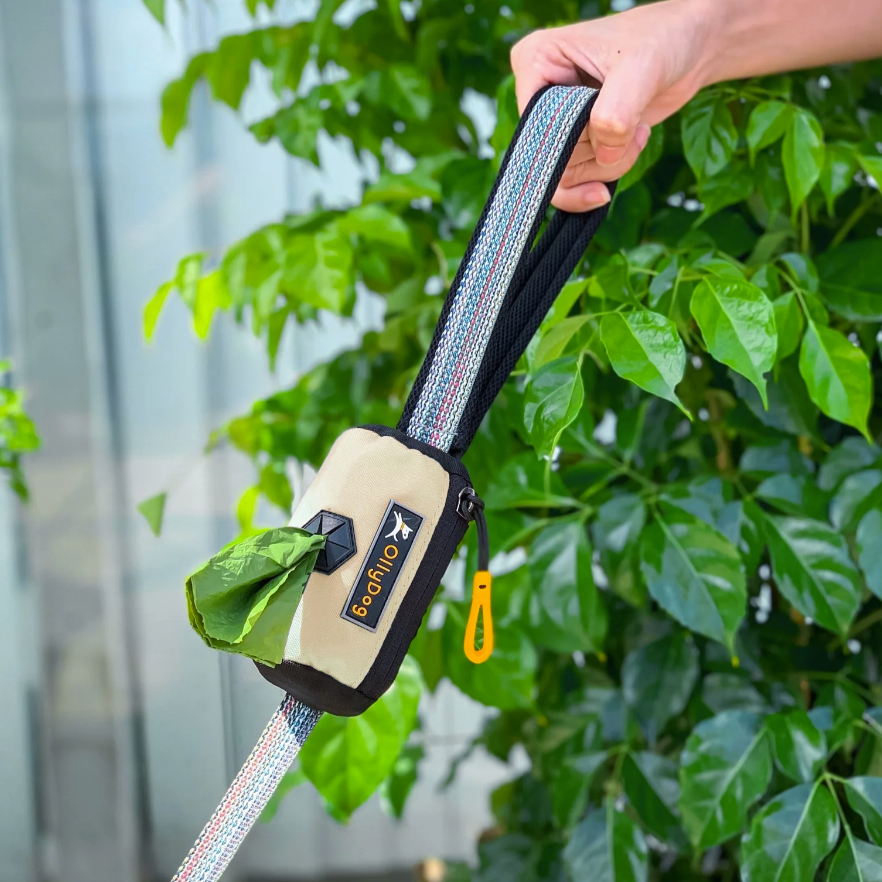 Close-up of a hand holding a dog leash with an attached doggie poop bag holder from OllyDog, Scoop Pick Up Bag in Bluff. #color_bluff