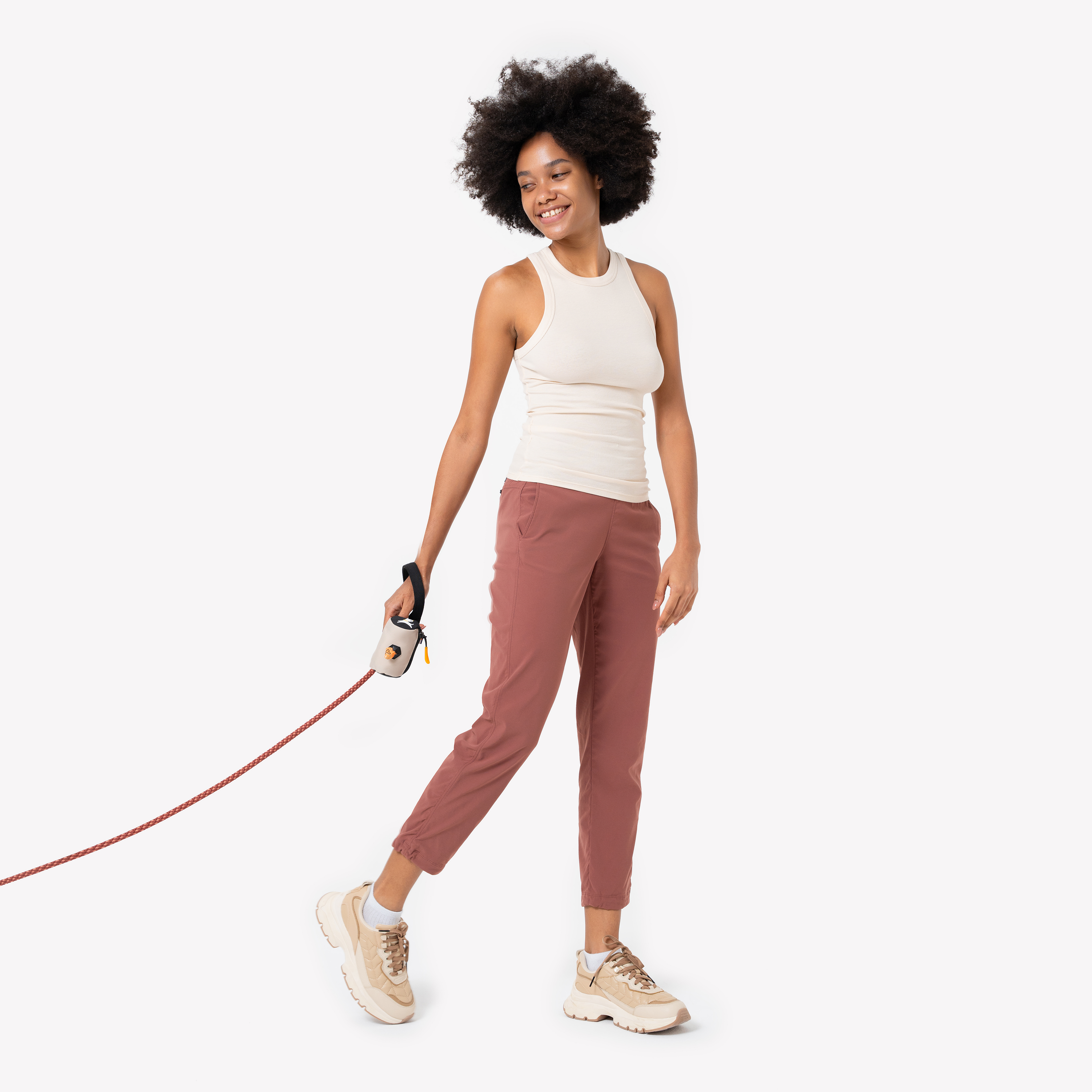 Full-length shot of a smiling woman with natural afro hair wearing a white sleeveless top, rust-colored pants, and beige sneakers, holding an orange dog leash with a beige-and-black waste-bag dispenser attached #color_con leche