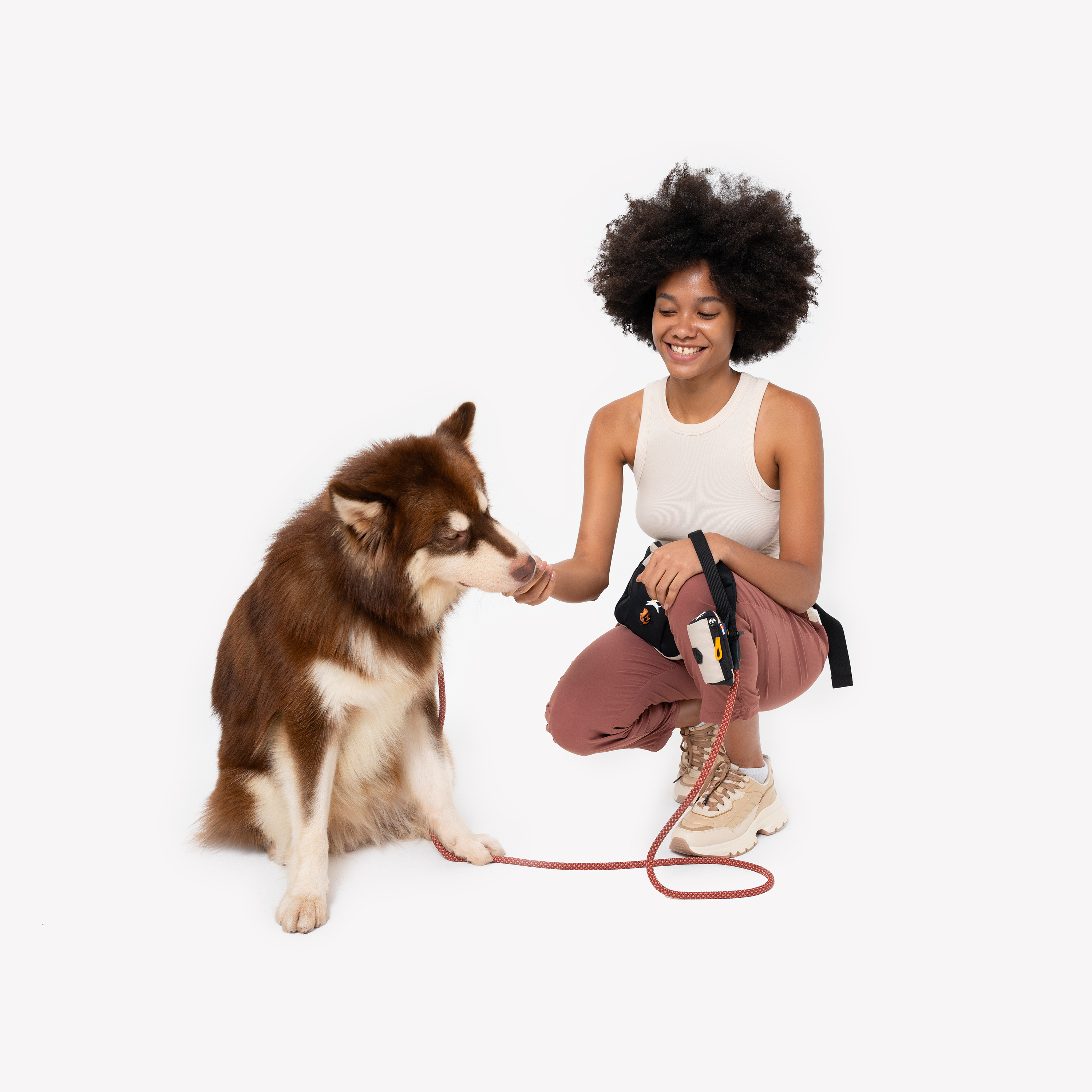 A smiling woman crouching on one knee in a white sleeveless top, rust-colored pants, and beige sneakers, holding an orange dog leash with a beige-and-black waste-bag dispenser attached, as she offers a treat to a brown-and-white husky #color_con leche