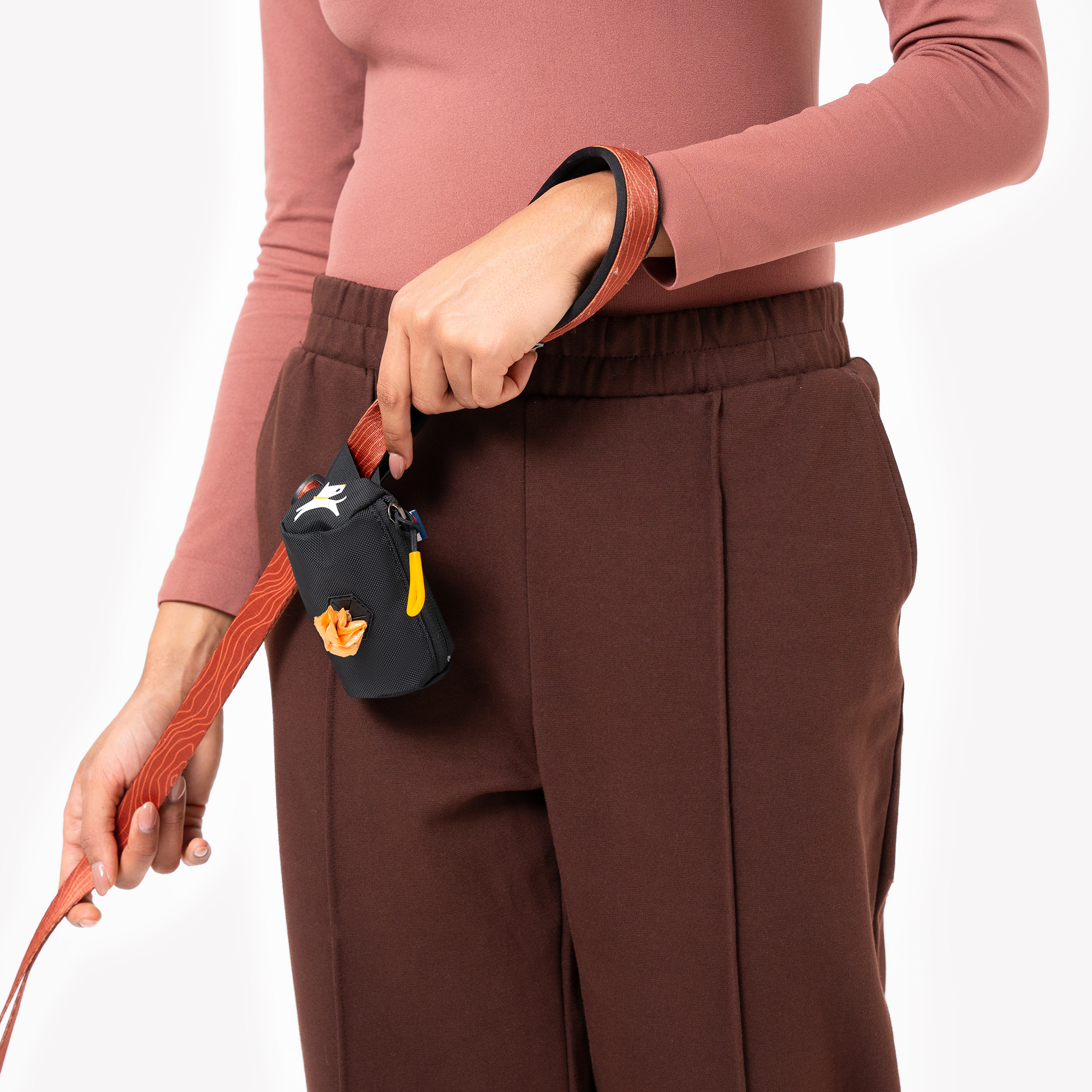 Person holding a dog leash with a black dog waste bag dispenser attached to their waistband, wearing brown pants and a pink long-sleeve top #color_raven