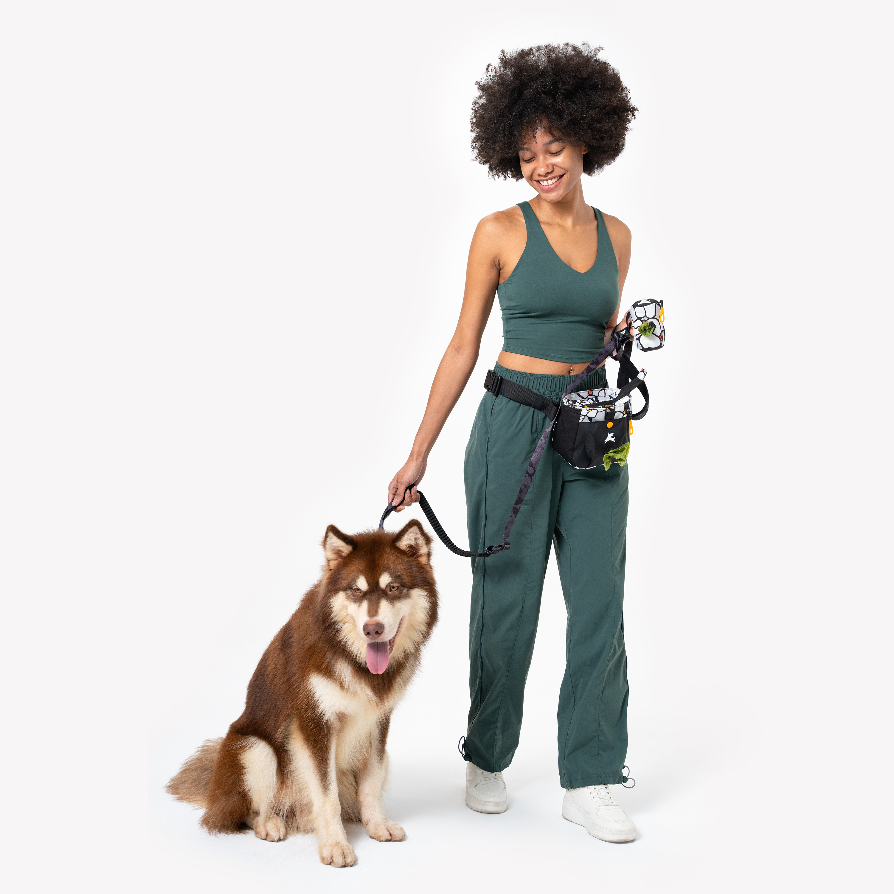 Woman walking a brown dog while carrying a patterned Scoop Pick Up Bag and a matching treat pouch attached to her waist, wearing a green athletic outfit #color_magnolia