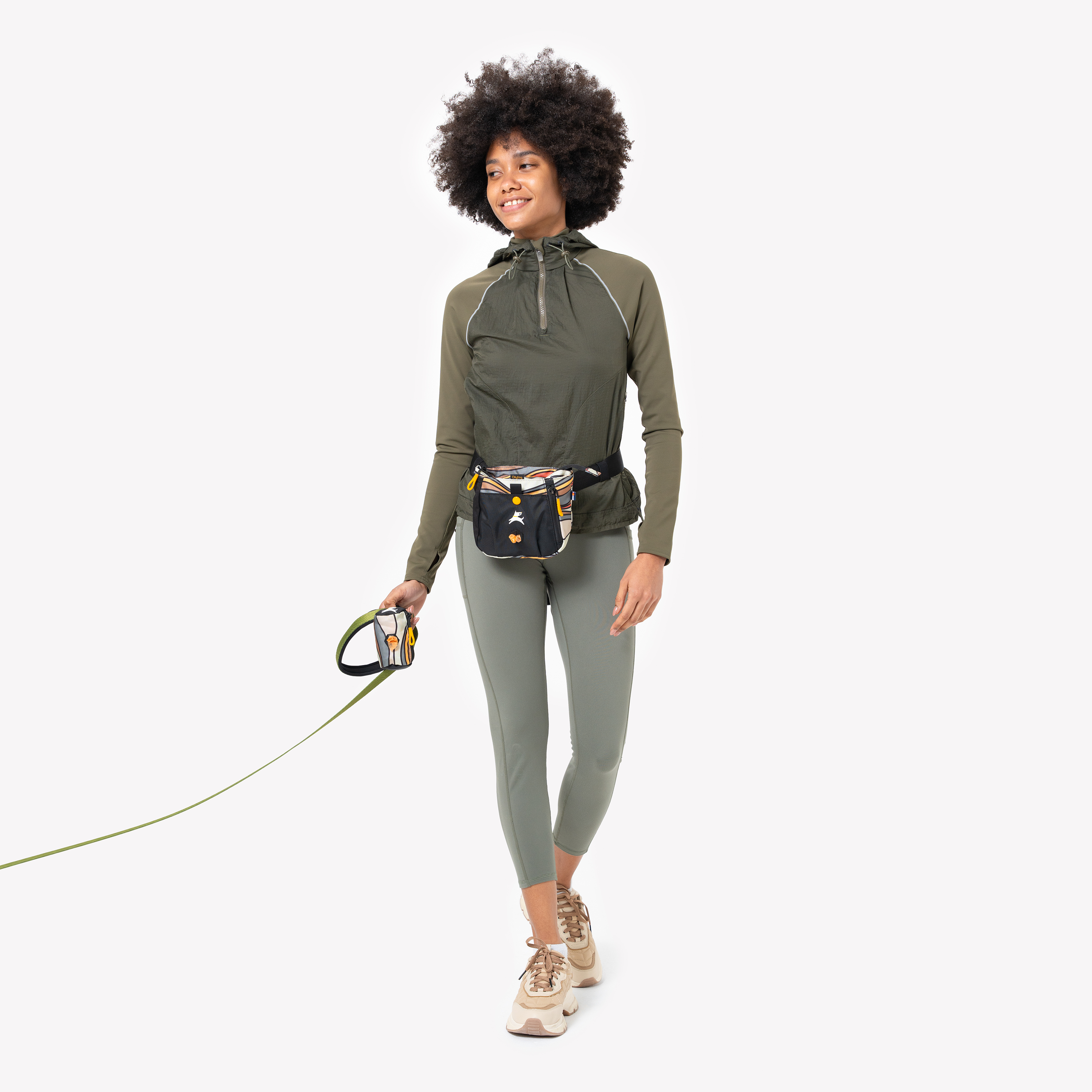 Woman walking with a green leash, carrying a patterned Scoop Pick Up Bag and matching treat pouch, dressed in green activewear and sneakers #color_ventura '73