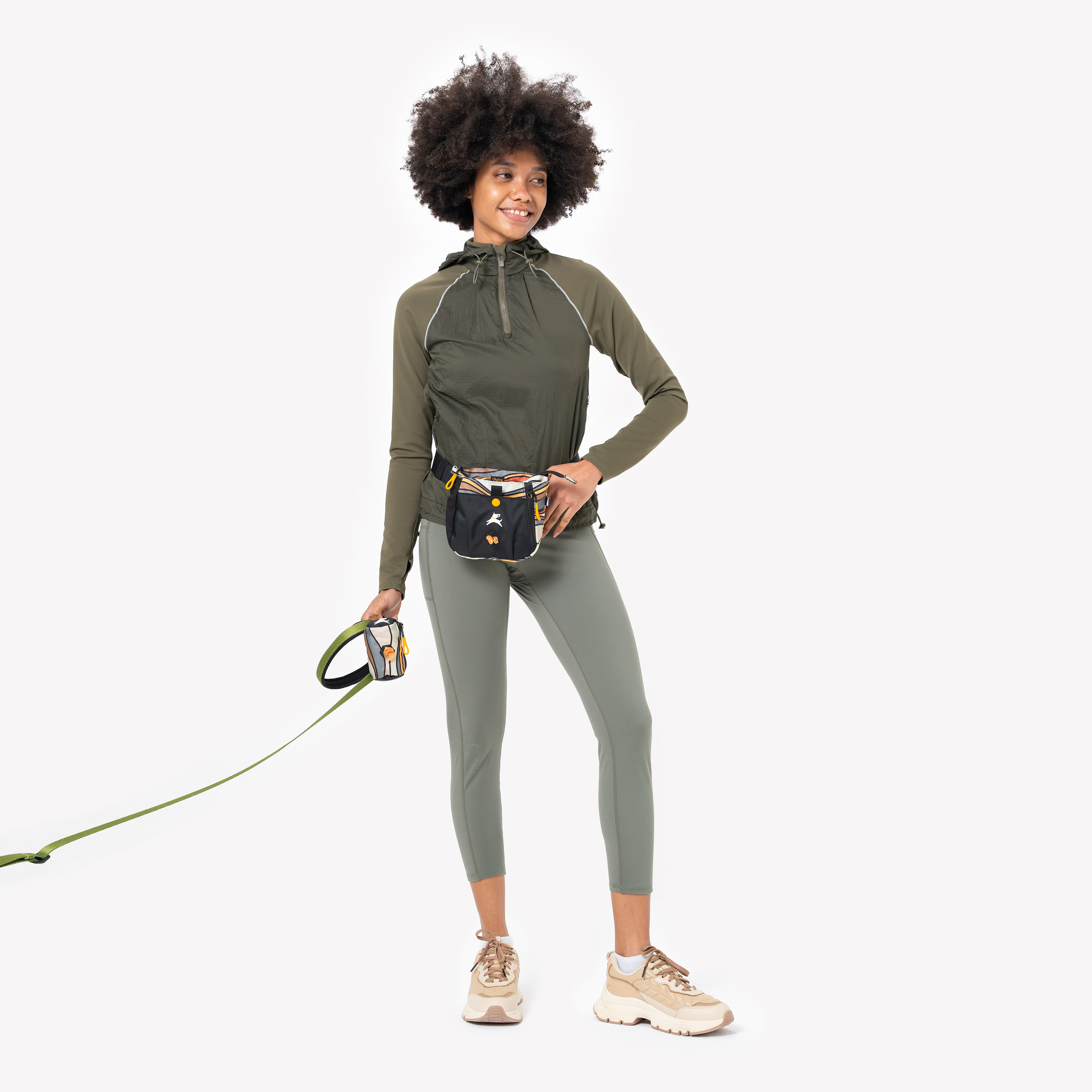 Woman holding a green leash with attached Scoop Pick Up Bag and wearing a matching treat pouch, dressed in green activewear and beige sneakers #color_ventura '73