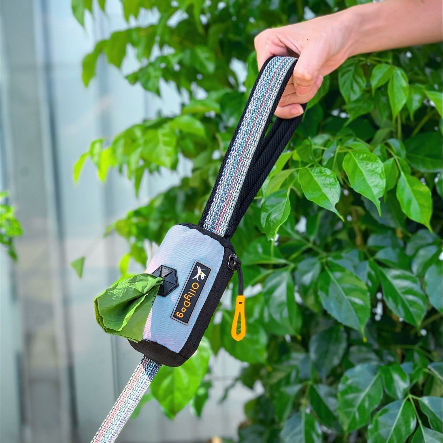 Close-up of a hand holding a dog leash with an attached doggie poop bag holder from OllyDog, Scoop Pick Up Bag in Maui Blue. #color_maui blue
