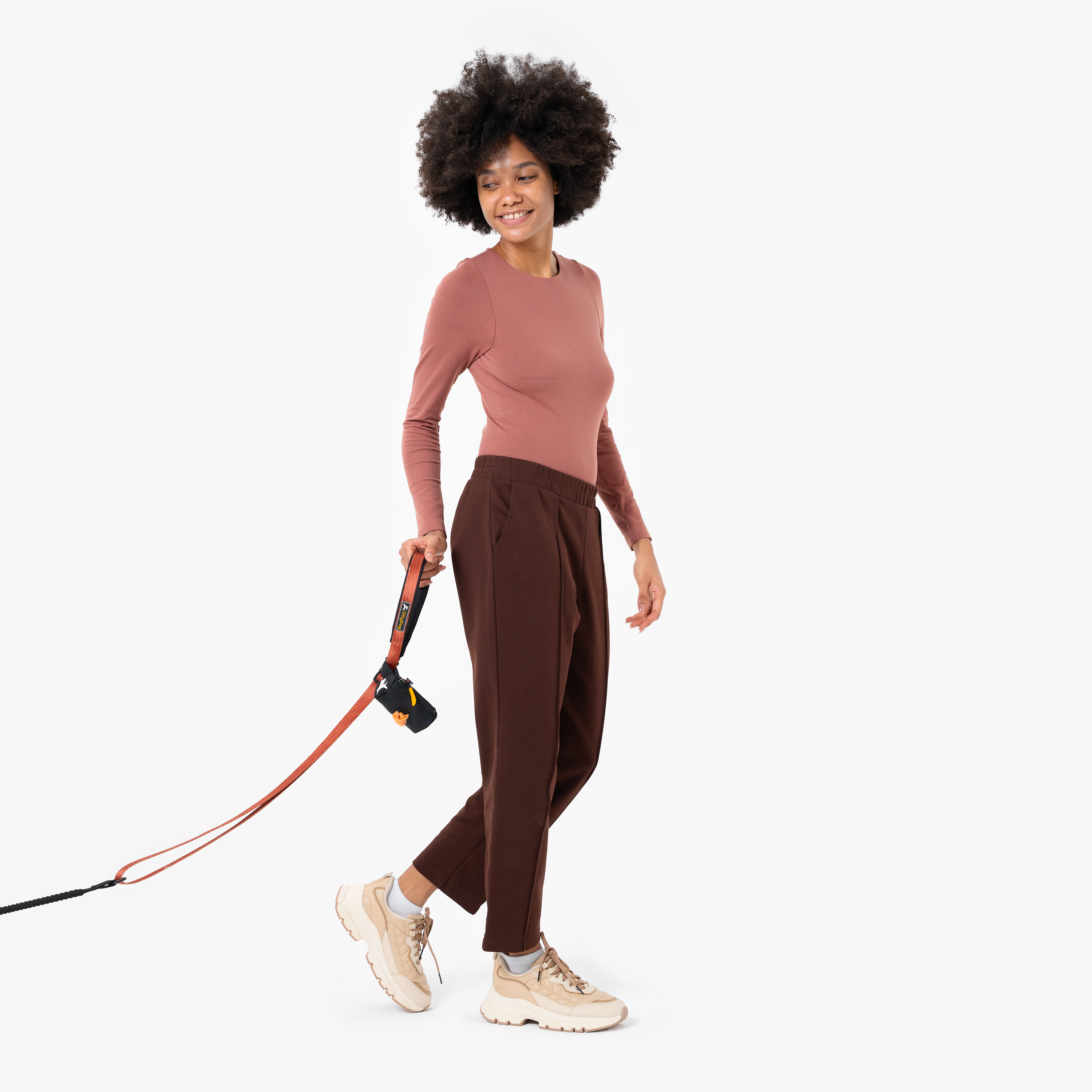 Full-length shot of a smiling woman with natural afro hair wearing a pink long-sleeve top, brown pants, and beige sneakers, holding an orange dog leash with a black waste-bag dispenser attached #color_raven
