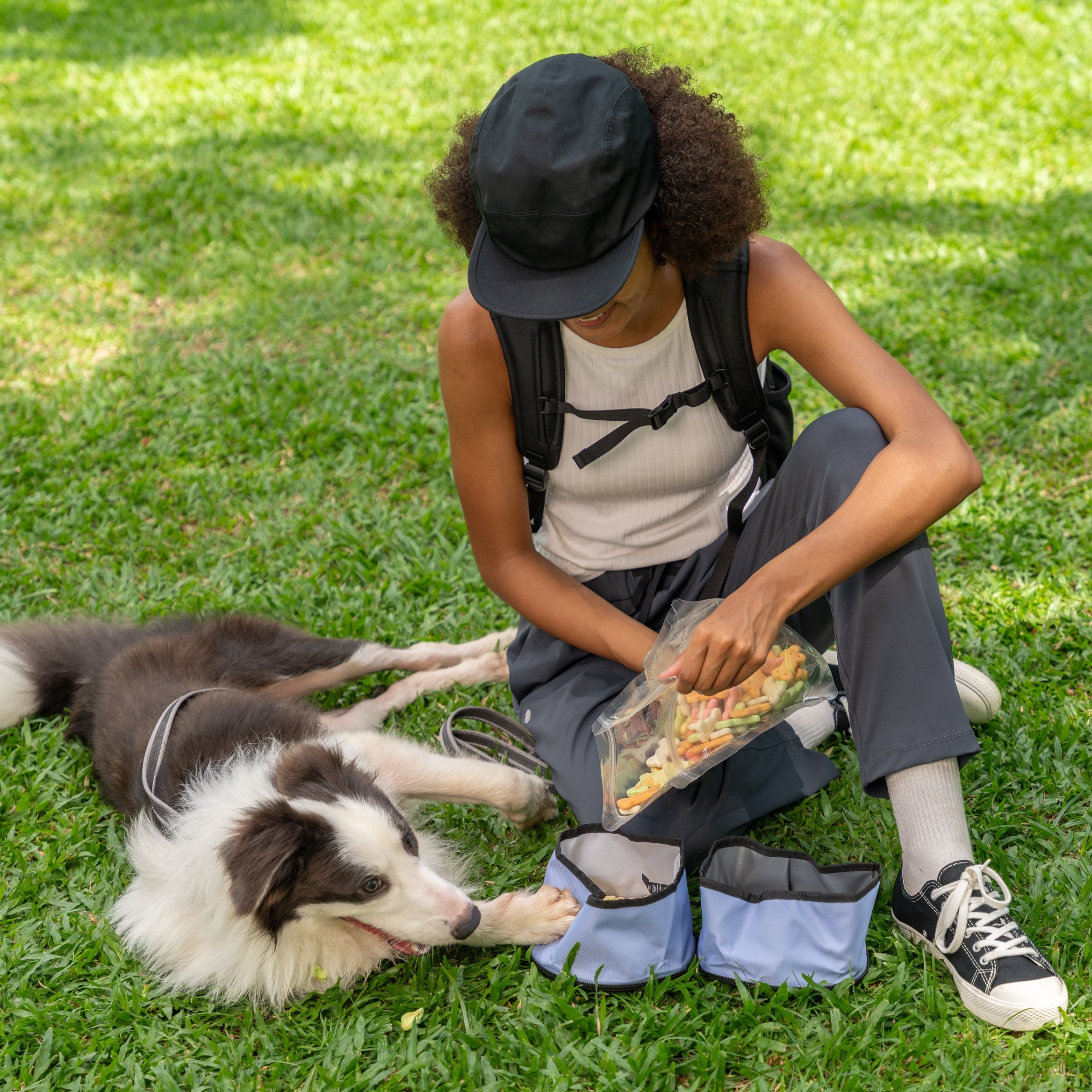 Woman sitting on grass with her dog while filling the OllyDog Superbowl in Blue Heron color with treats, featuring dual recycled bowls for food and water #color_blue heron