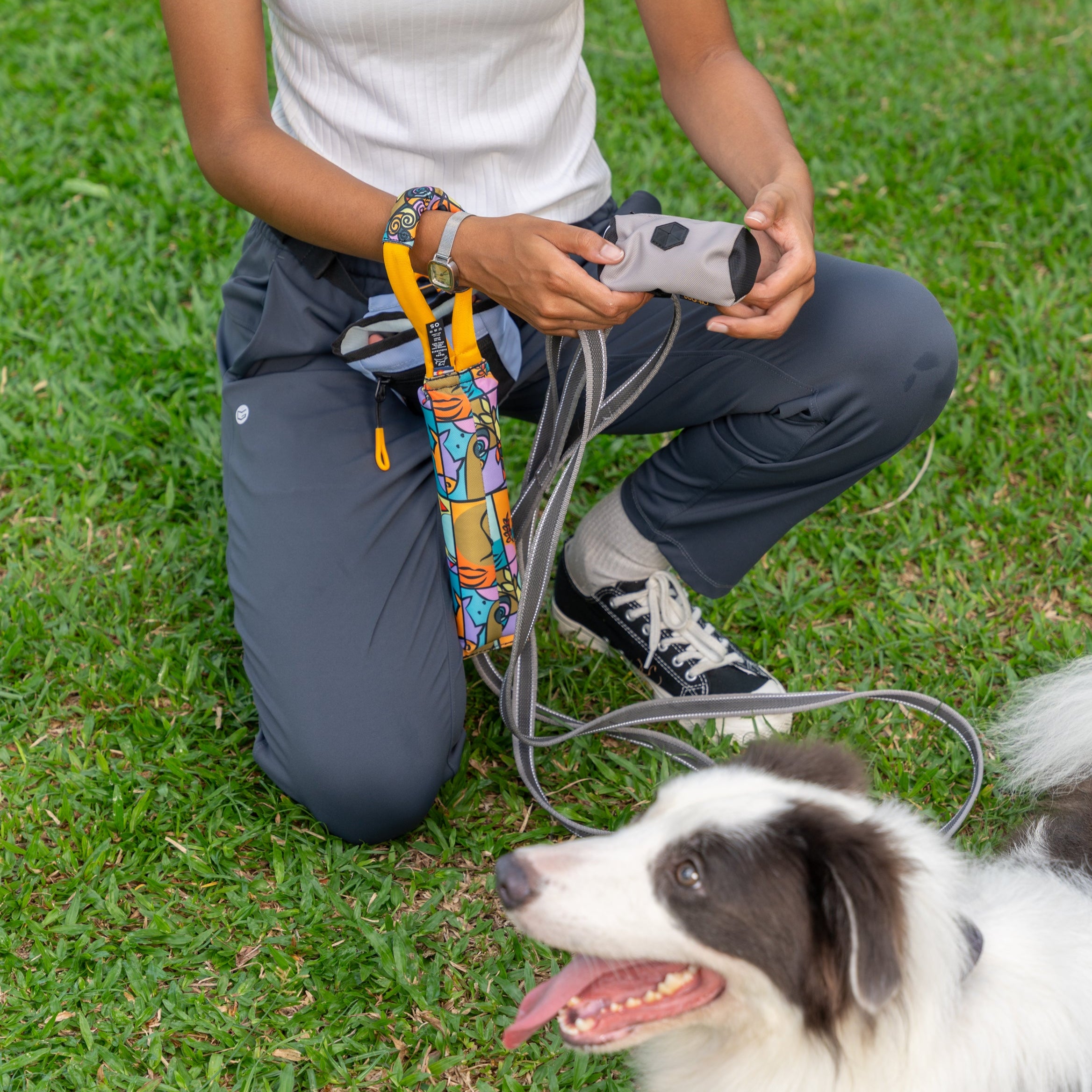Woman kneeling in the grass with her dog, showing the OllyDog Tugger Toy in Cloud9 pattern and Scoop Pick Up Bag in Bluff color attached to her waist #color_cloud9