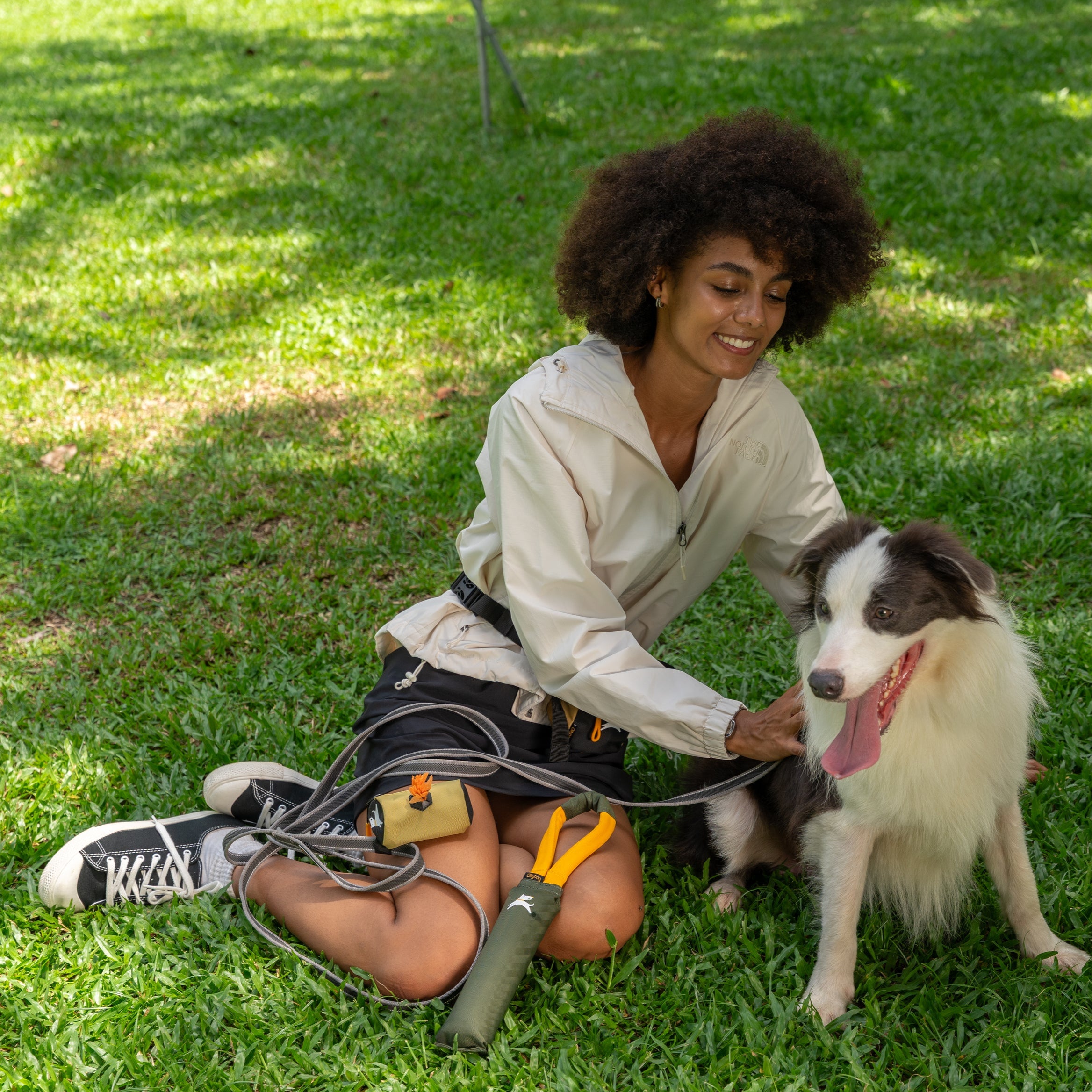 Woman sitting on the grass with her dog, showing the OllyDog Tugger Toy in Winter Moss and Scoop Pick Up Bag in Alchemy color during a sunny park outing #color_winter moss