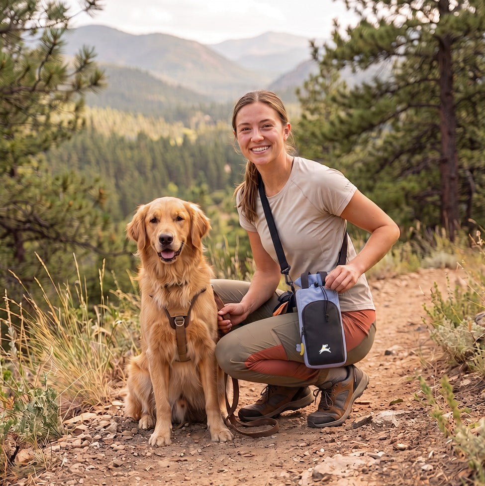 Lifestyle photo of a woman kneeling on a mountain dirt trail with her golden retriever, wearing the OllyDog OllyCaddy dog walking carryall in Blue Heron color across her body.
#color_blue heron