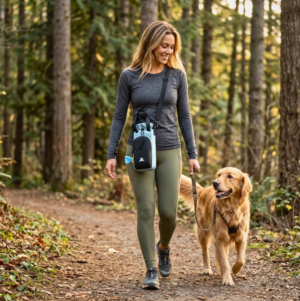 Lifestyle photo of a woman walking a golden retriever on a forest trail while wearing the OllyDog OllyCaddy dog walking carryall in St. Lucia color across her body for hands-free convenience. #color_st. lucia