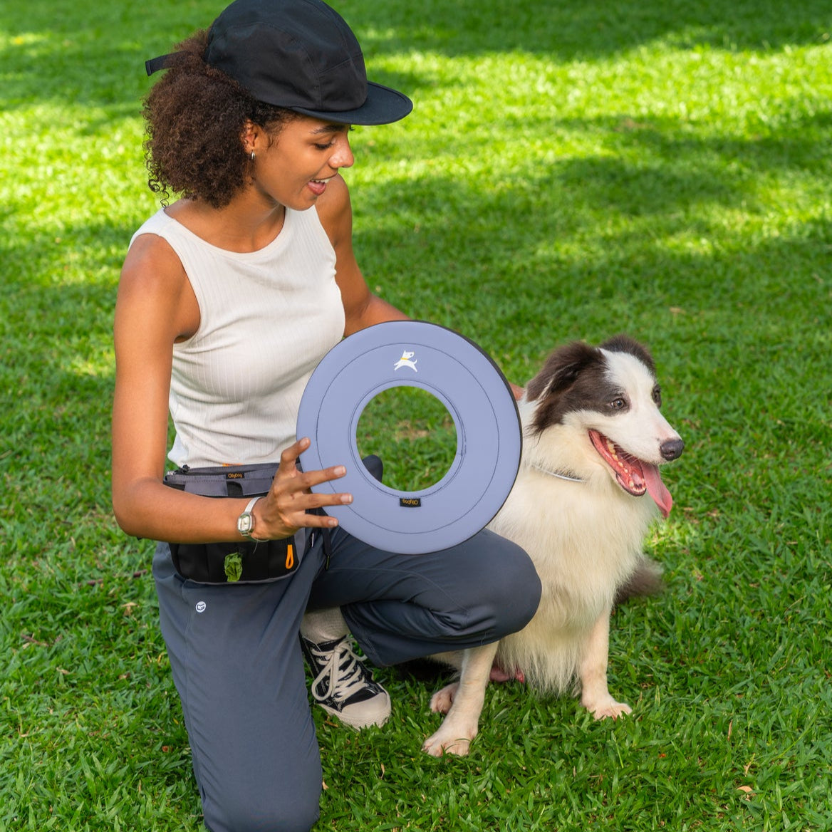 Lifestyle shot of a smiling woman holding the OllyDog Super Flyer dog flying disc in blue heron color next to a happy black and white border collie on a green lawn.#color_blue heron