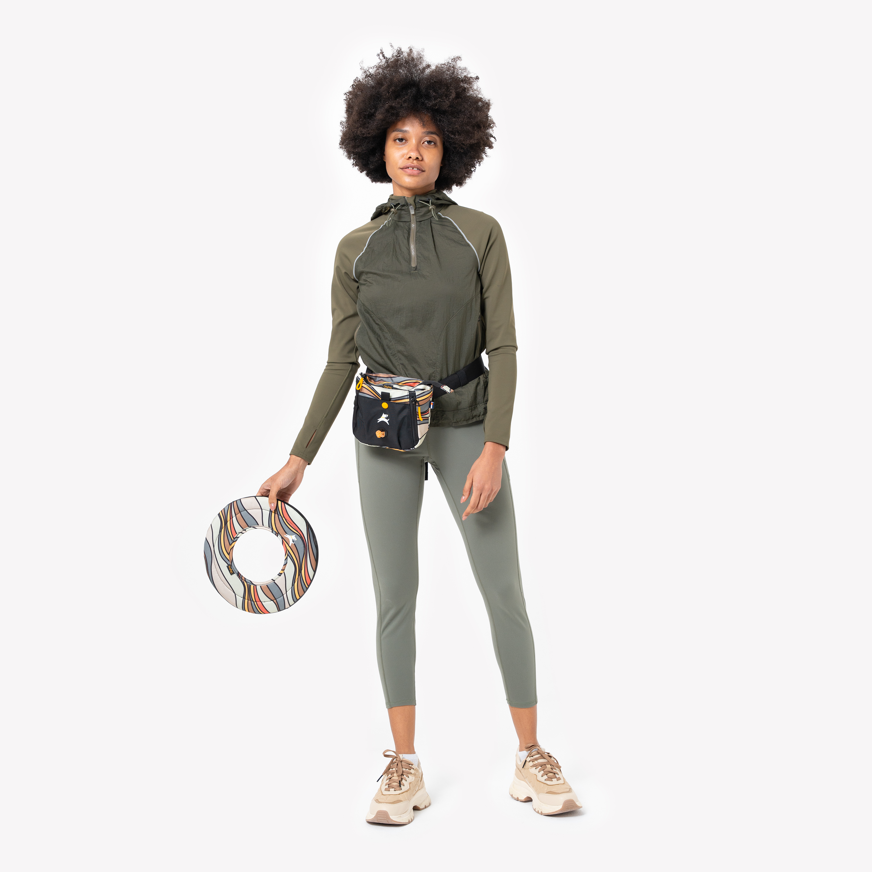 Woman in green activewear holding Ventura '73 Super Flyer dog frisbee with retro wave pattern, wearing matching treat pouch and beige sneakers #color_ventura '73