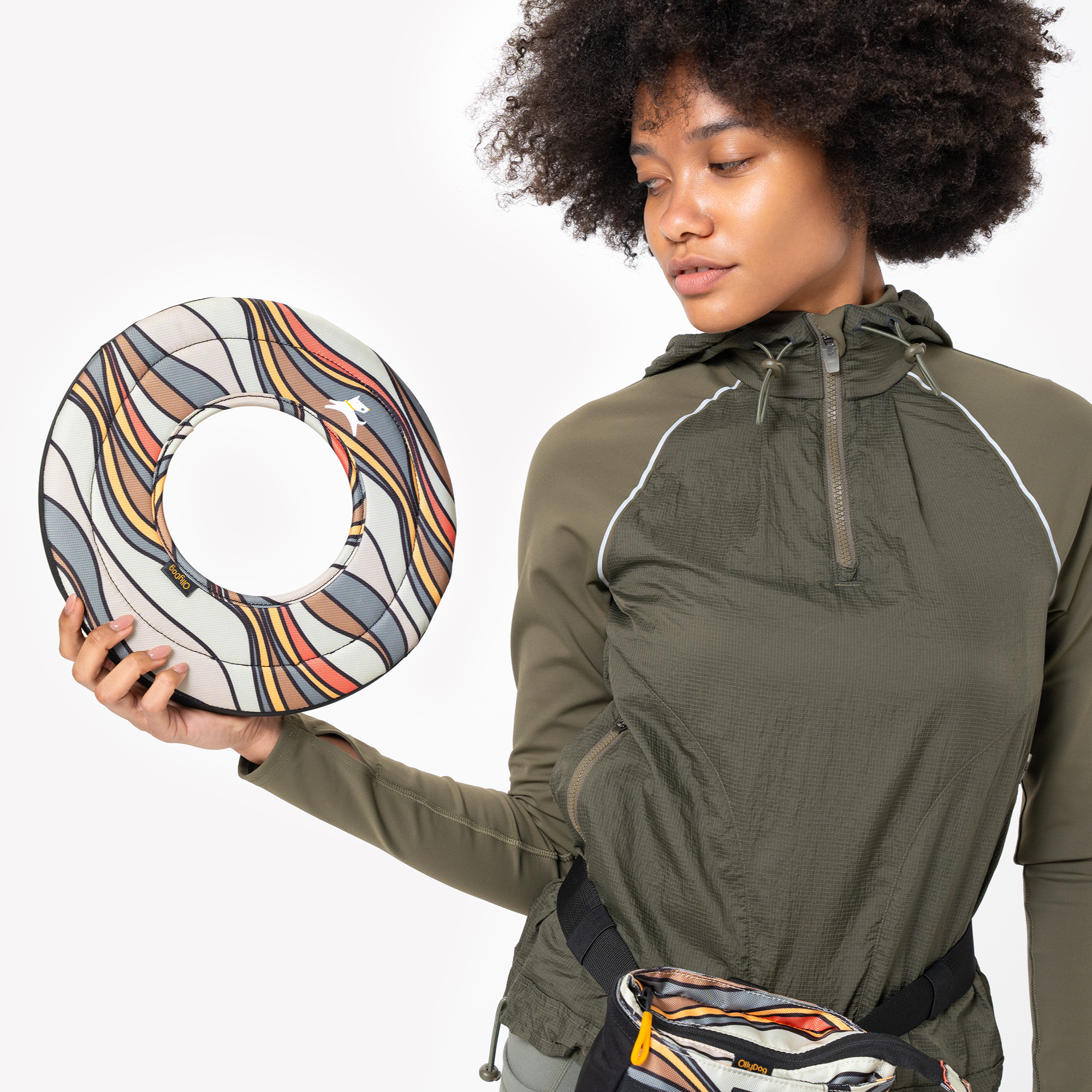 Close-up of woman in green jacket holding Ventura '73 Super Flyer fabric dog frisbee with retro wave pattern, wearing matching treat pouch #color_ventura '73