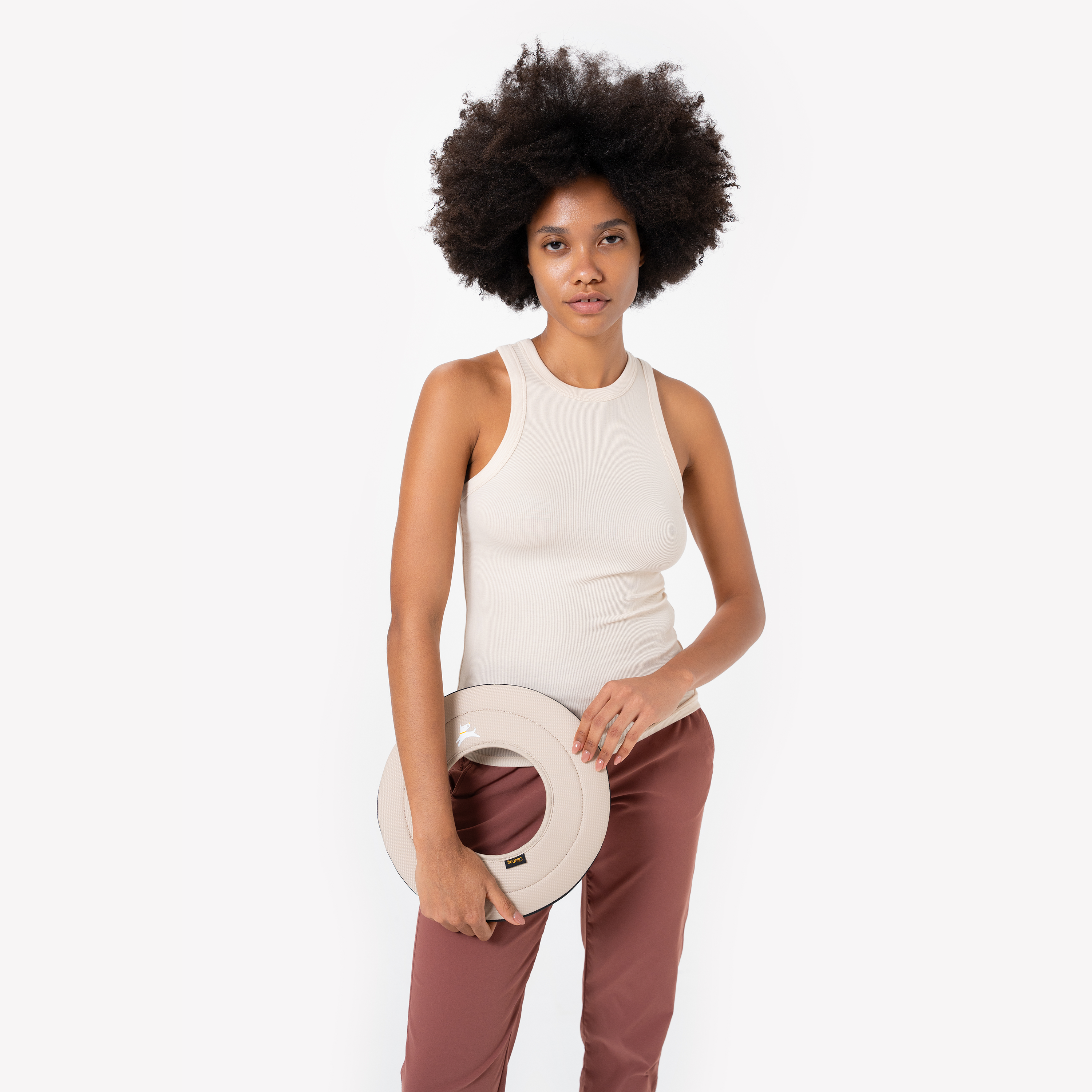 Woman in a cream tank top and burgundy pants holding a beige Super Flyer fabric dog frisbee in Con Leche color #color_con leche