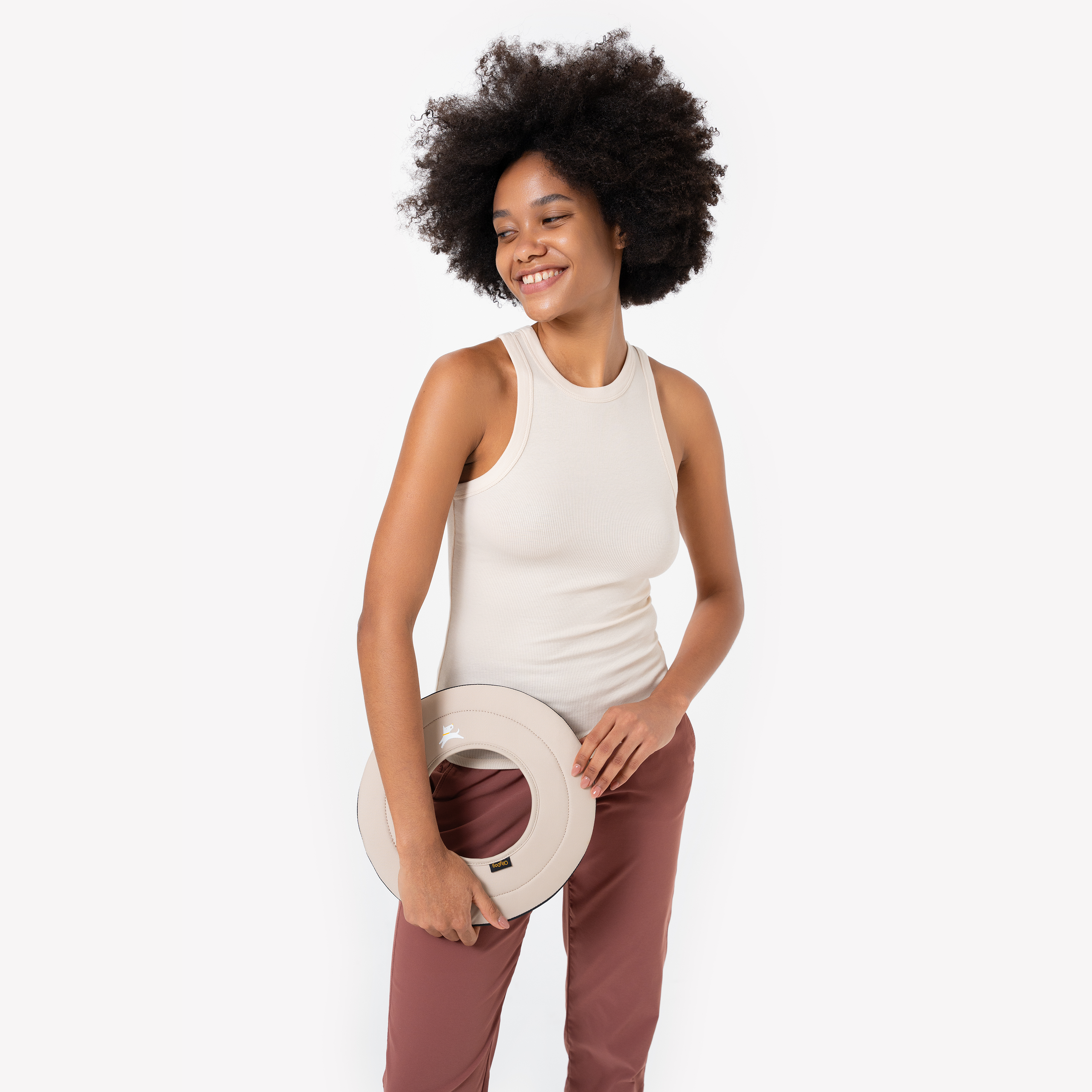 Woman in a beige tank top and maroon pants holding the Super Flyer fabric dog frisbee in Con Leche color by her side, smiling #color_con leche