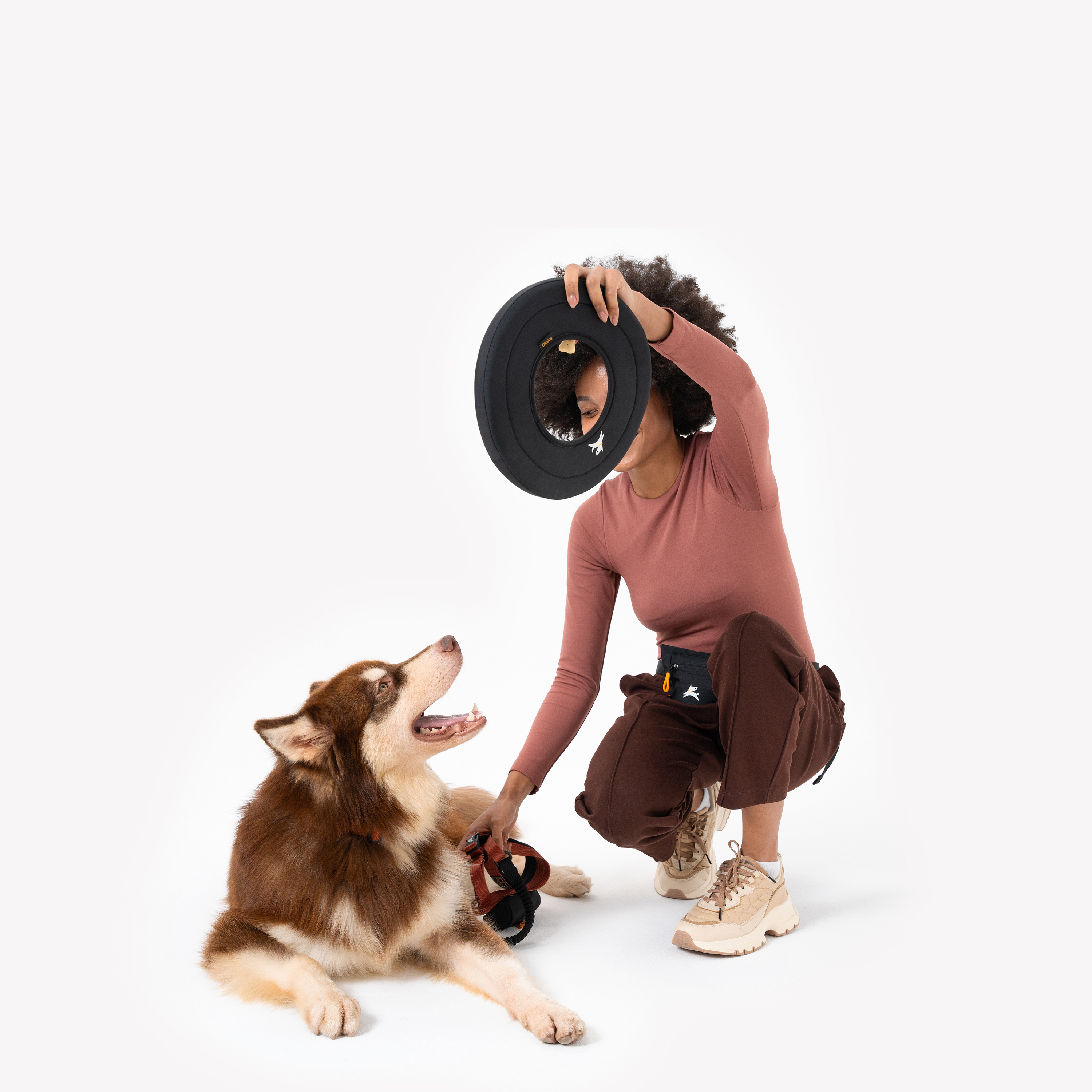 Woman squatting and holding a black OllyDog Super Flyer frisbee above her head while playing with a brown and white husky dog, color Raven. #color_raven