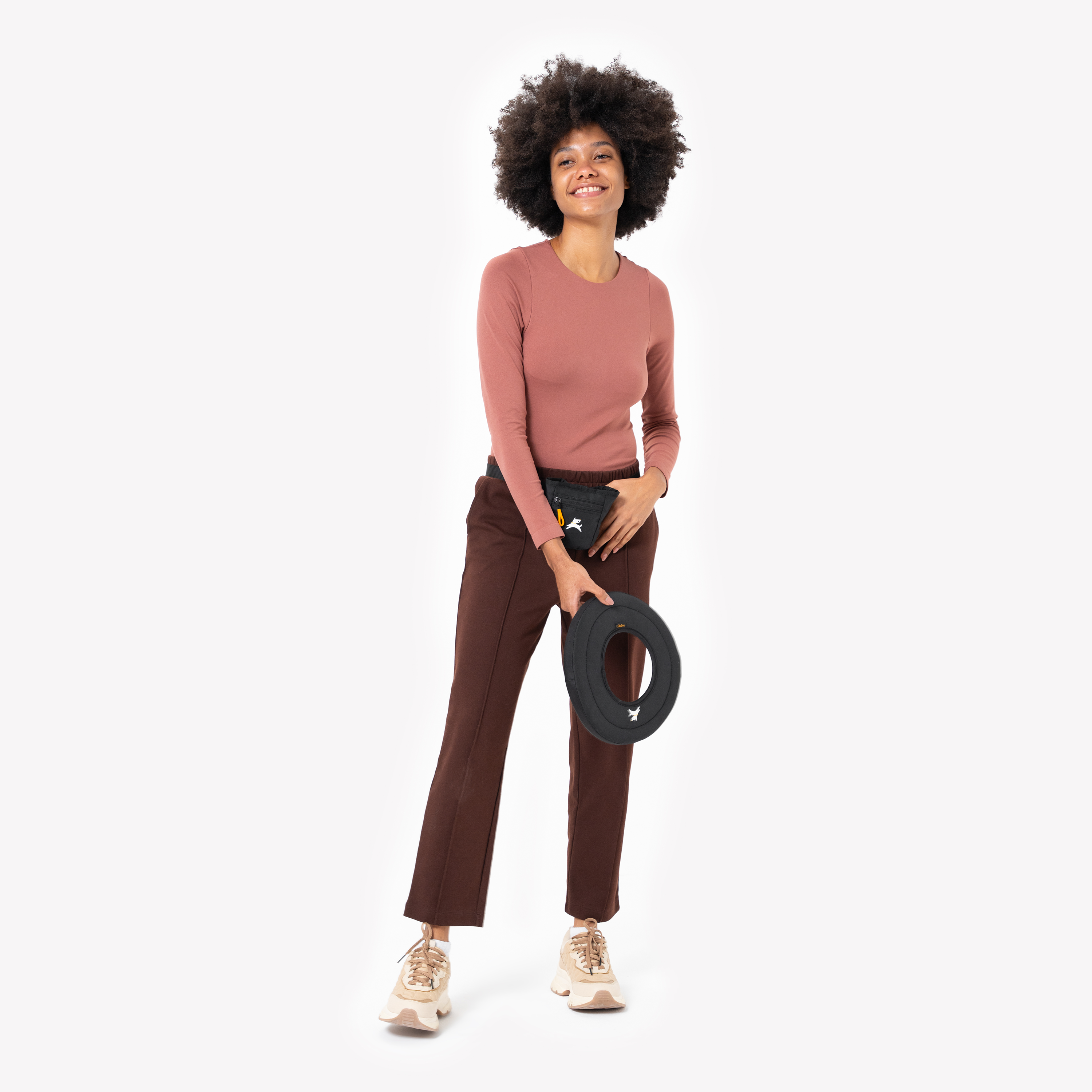 Woman in a rose long-sleeve top and brown pants holding a black OllyDog Super Flyer fabric dog frisbee, color Raven, while smiling and standing against a white background #color_raven