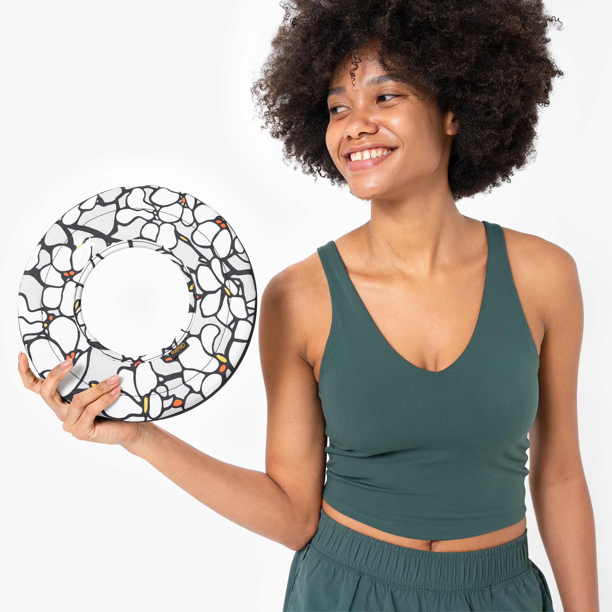 Woman in green workout top smiling while holding a Magnolia Super Flyer dog frisbee with black and white floral pattern and orange accents. #color_magnolia