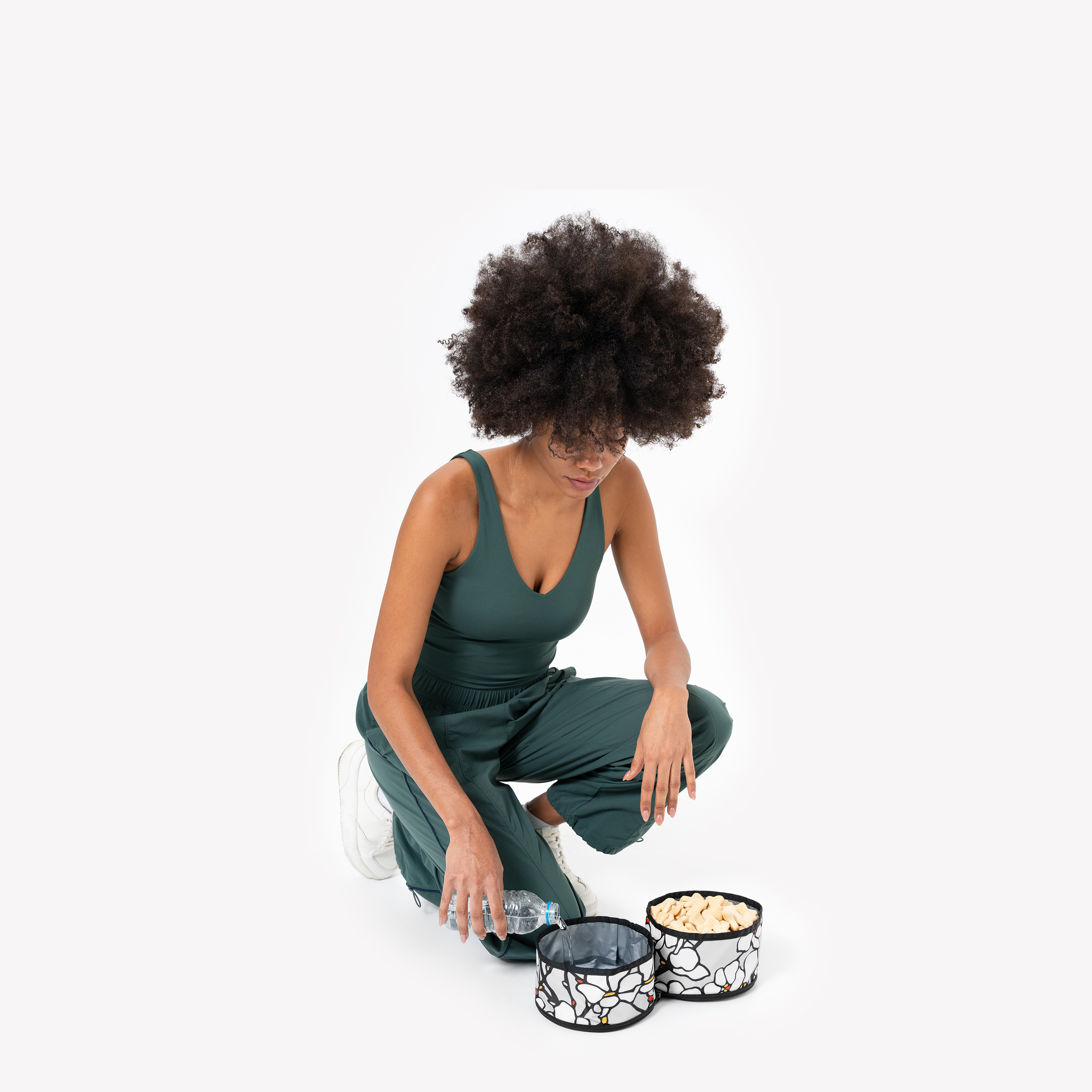 A person with dark, voluminous curly hair, wearing a dark green jumpsuit, is crouched and pouring water from a clear container into one of two Magnolia collapsible dog bowls featuring a black and white floral pattern. The other bowl contains yellow kibble, set against a white background #color_magnolia