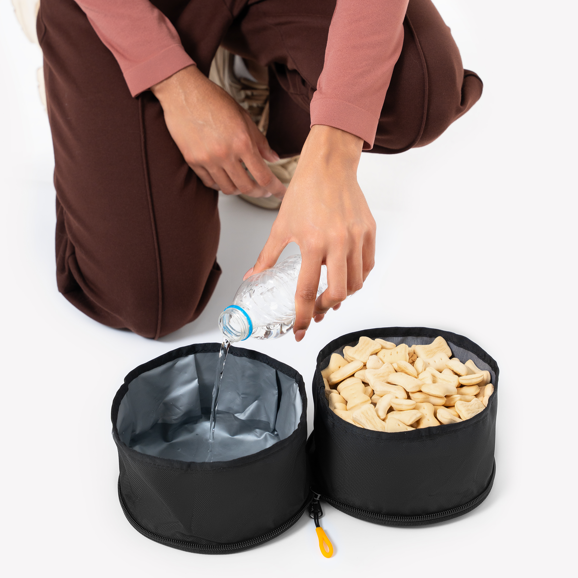 A person is pouring water from a clear bottle into one compartment of a collapsible dog bowl made of black fabric. The other compartment contains light-colored kibble #color_raven