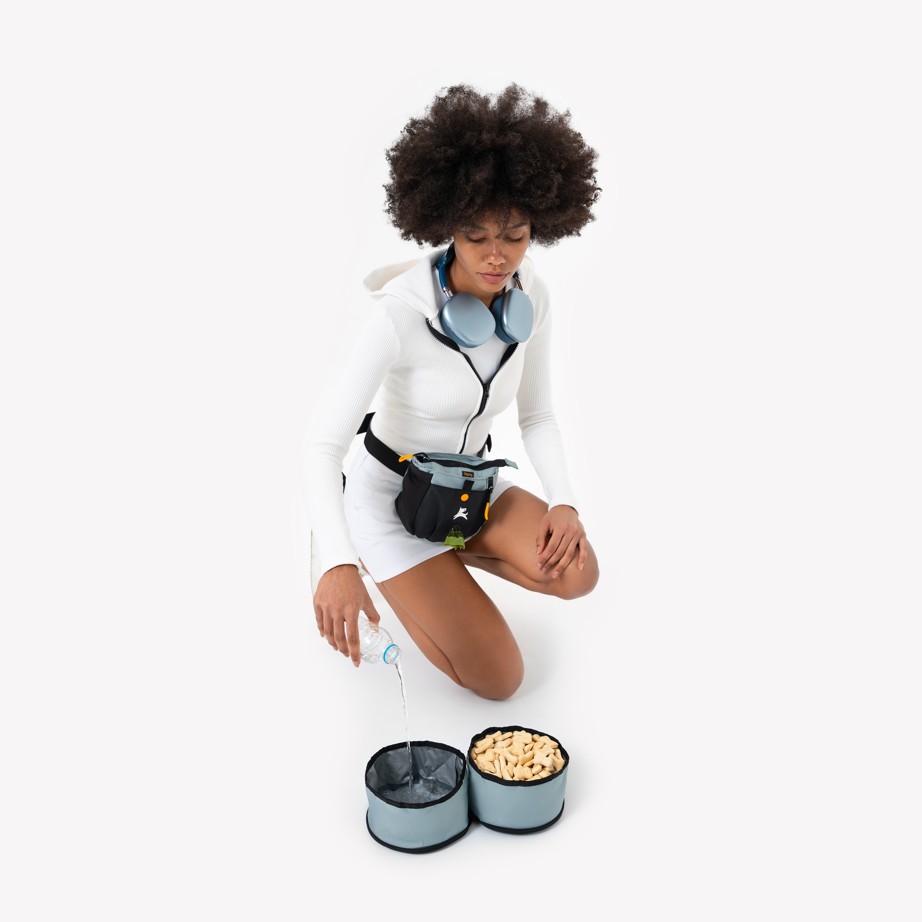 A person with dark, voluminous curly hair, wearing a white athletic outfit and a light blue fanny pack, is kneeling on a white background next to two light blue OllyDog Superbowl collapsible dog bowls. One bowl contain water, and the other holds yellow kibble #color_st. lucia