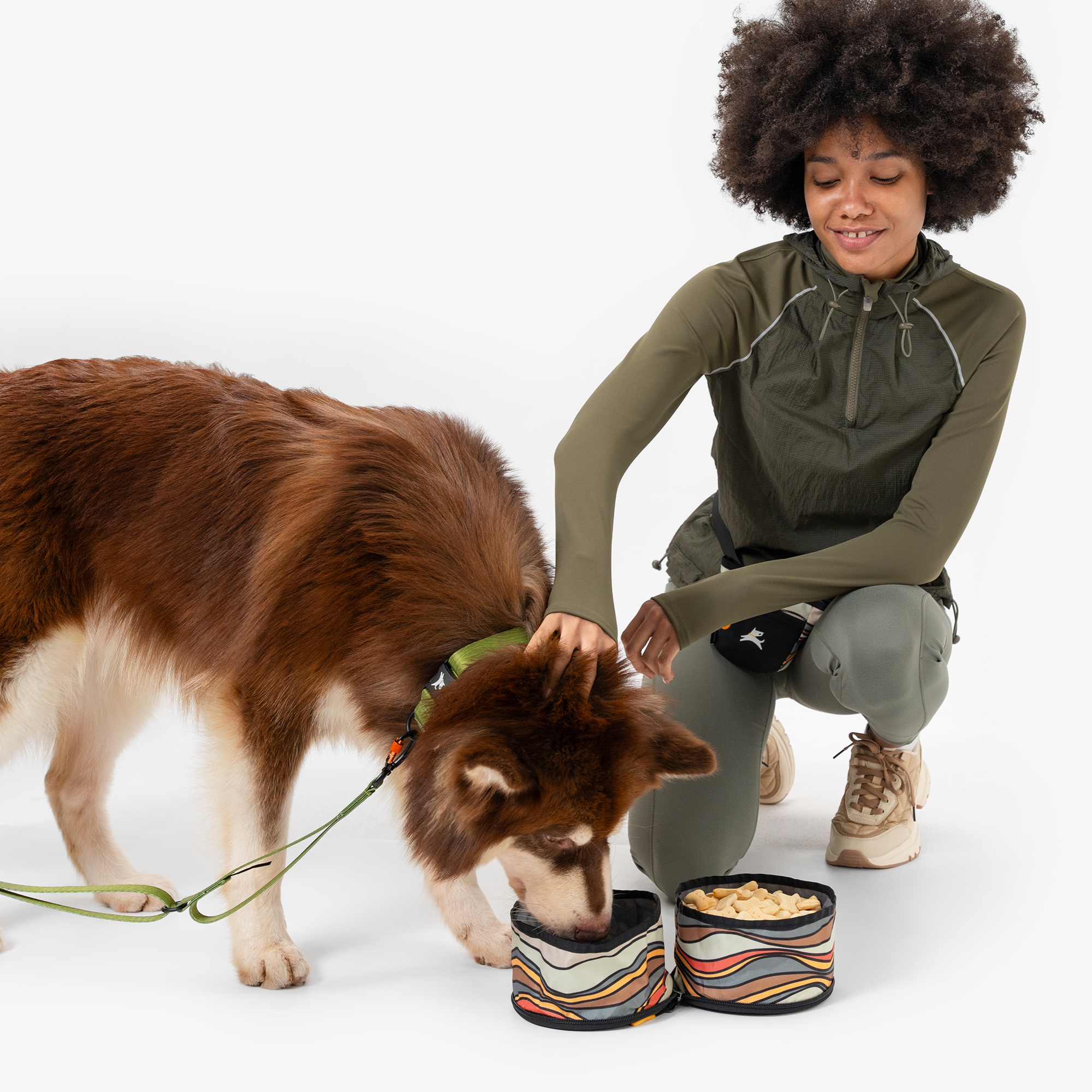 A person with dark curly hair, wearing an olive green jacket and gray pants, is kneeling next to a brown and white dog that is eating from one of two connected Ventura '73 collapsible dog bowls featuring a retro horizontal stripe pattern. The person is holding the dog's leash #color_ventura '73