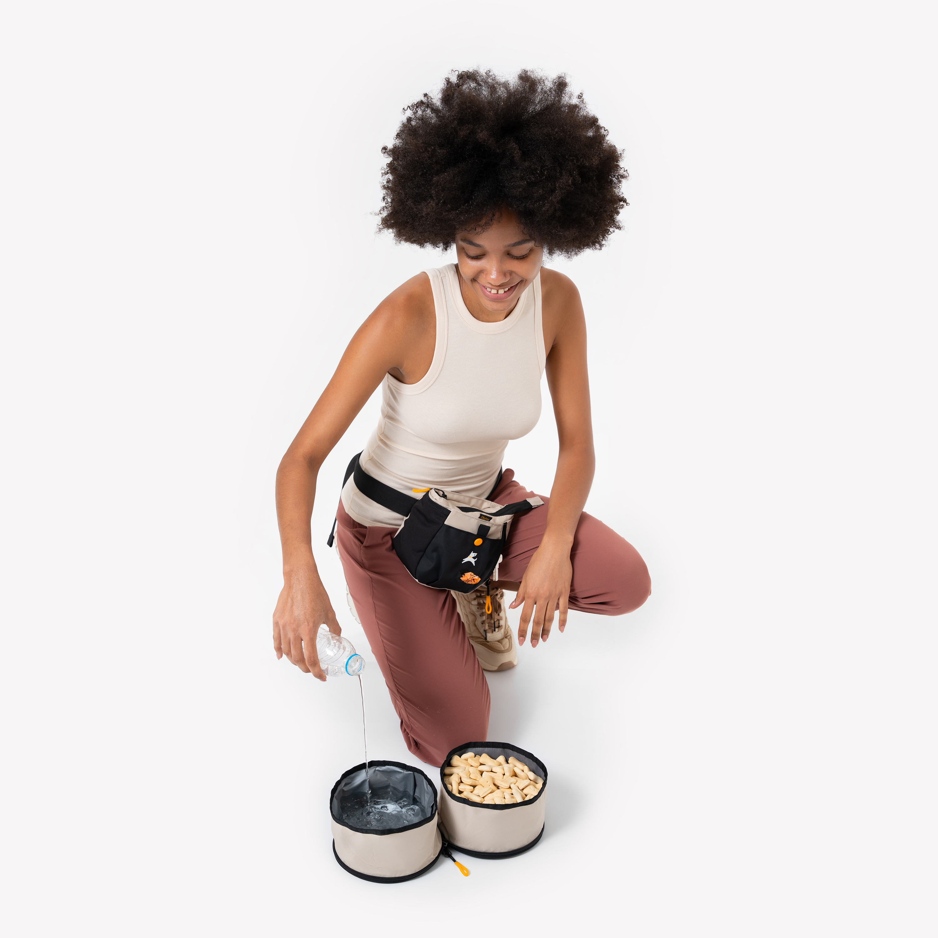 Woman in a cream tank top and mauve pants pouring water into the OllyDog Superbowl, which has two collapsible compartments for food and water, color Con Leche, shown in use with treats and hydration setup #color_con leche