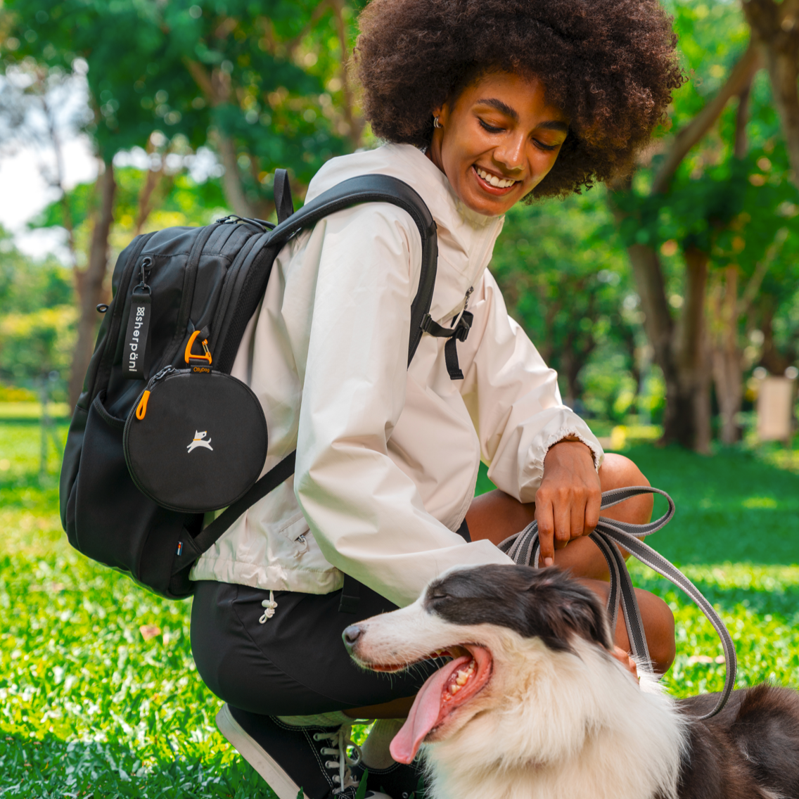Smiling woman kneeling in the park with the black backpack and attached Superbowl collapsible dog bowl, holding leash of happy black and white dog – ideal outdoor setup for pet owners and dog walkers #color_con leche
