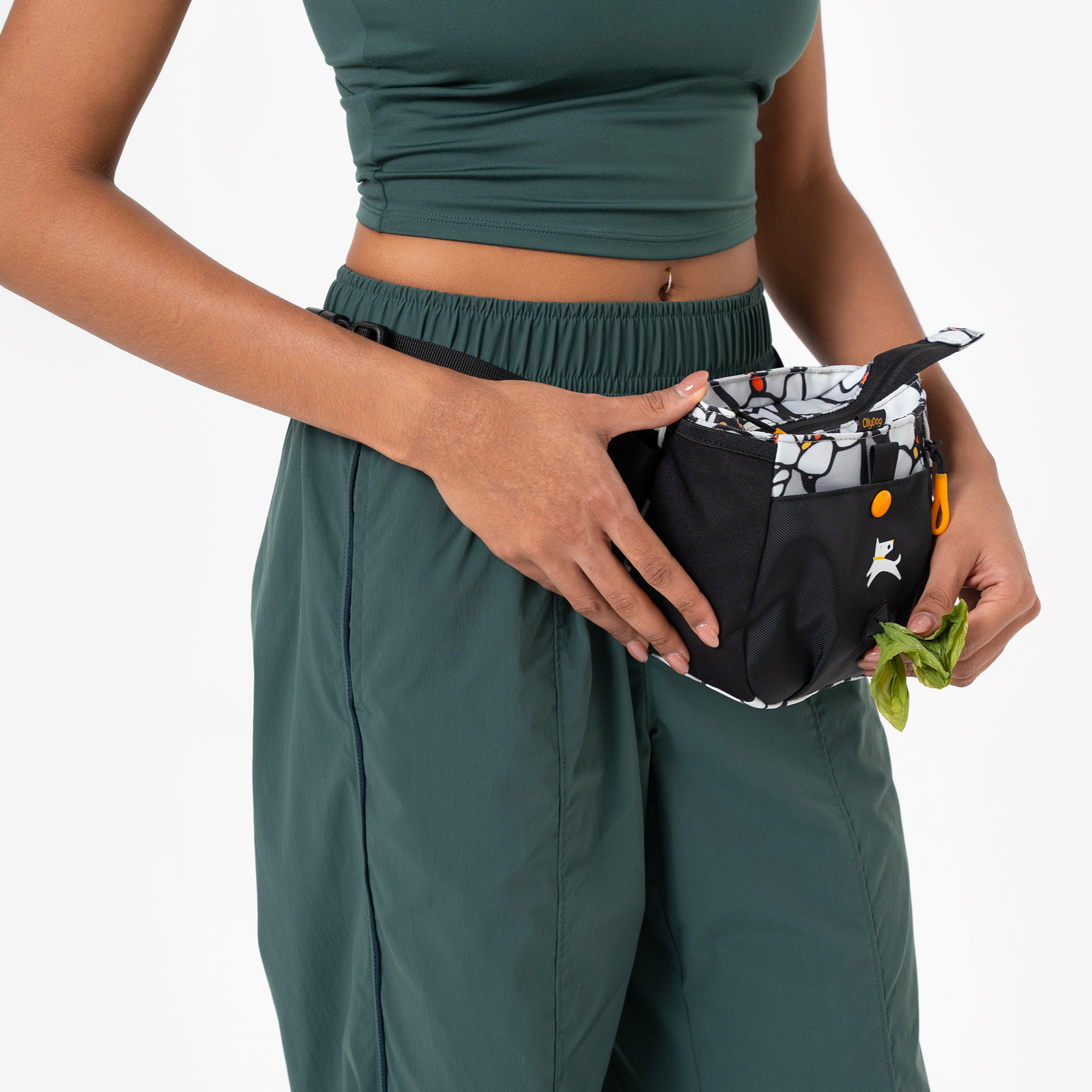 Person wearing a green crop top and pants, holding a black and patterned gardening belt pouch with fresh herbs inside #color_magnolia