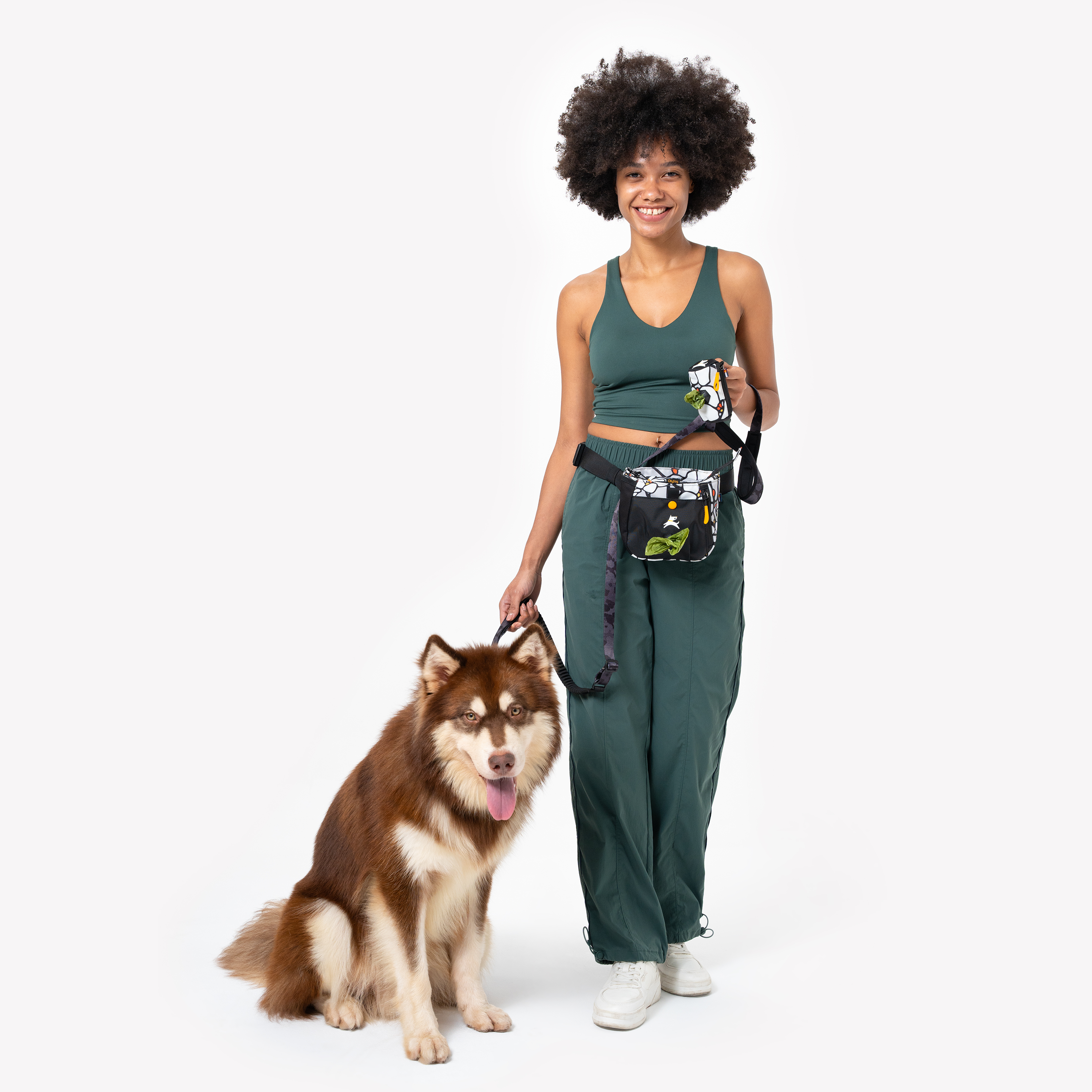 Smiling woman in green activewear holding a dog leash, wearing a patterned dog walking belt pouch, standing next to a fluffy brown and white dog #color_magnolia