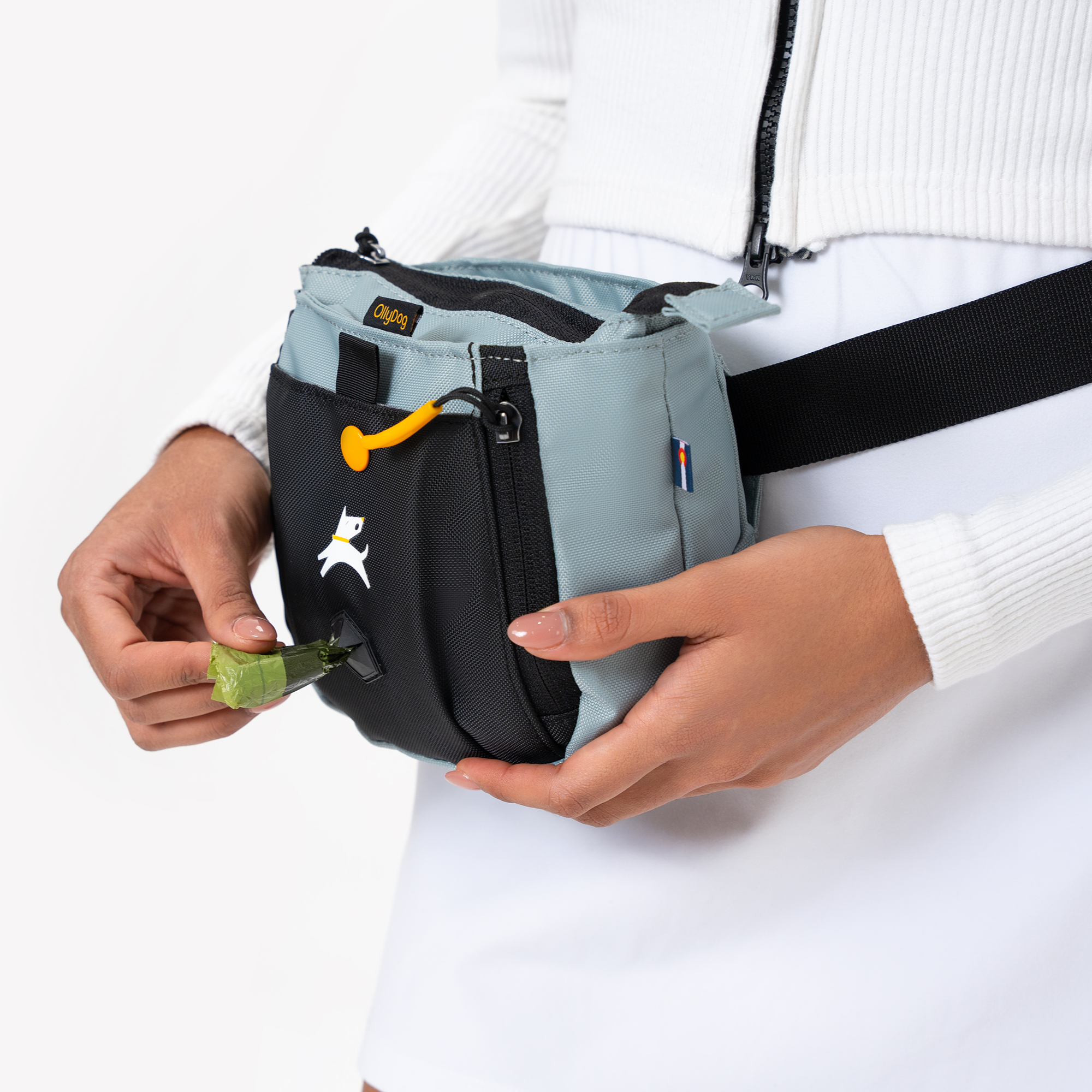 Close-up of a woman pulling a green waste bag from the built-in dispenser of a two-tone blue and black OllyDog waist bag, wearing a white zip-up top and skirt #color_st. lucia