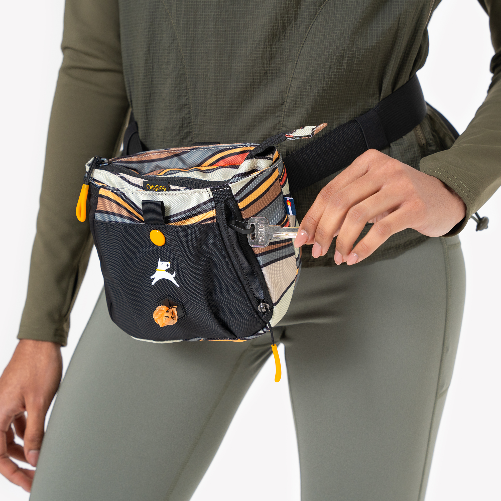 Close-up of a person in olive green athletic wear wearing a black and orange swirl-patterned dog walking waist pouch, placing a key into the zippered side pocket with a waste bag dispenser visible at the front #color_ventura '73