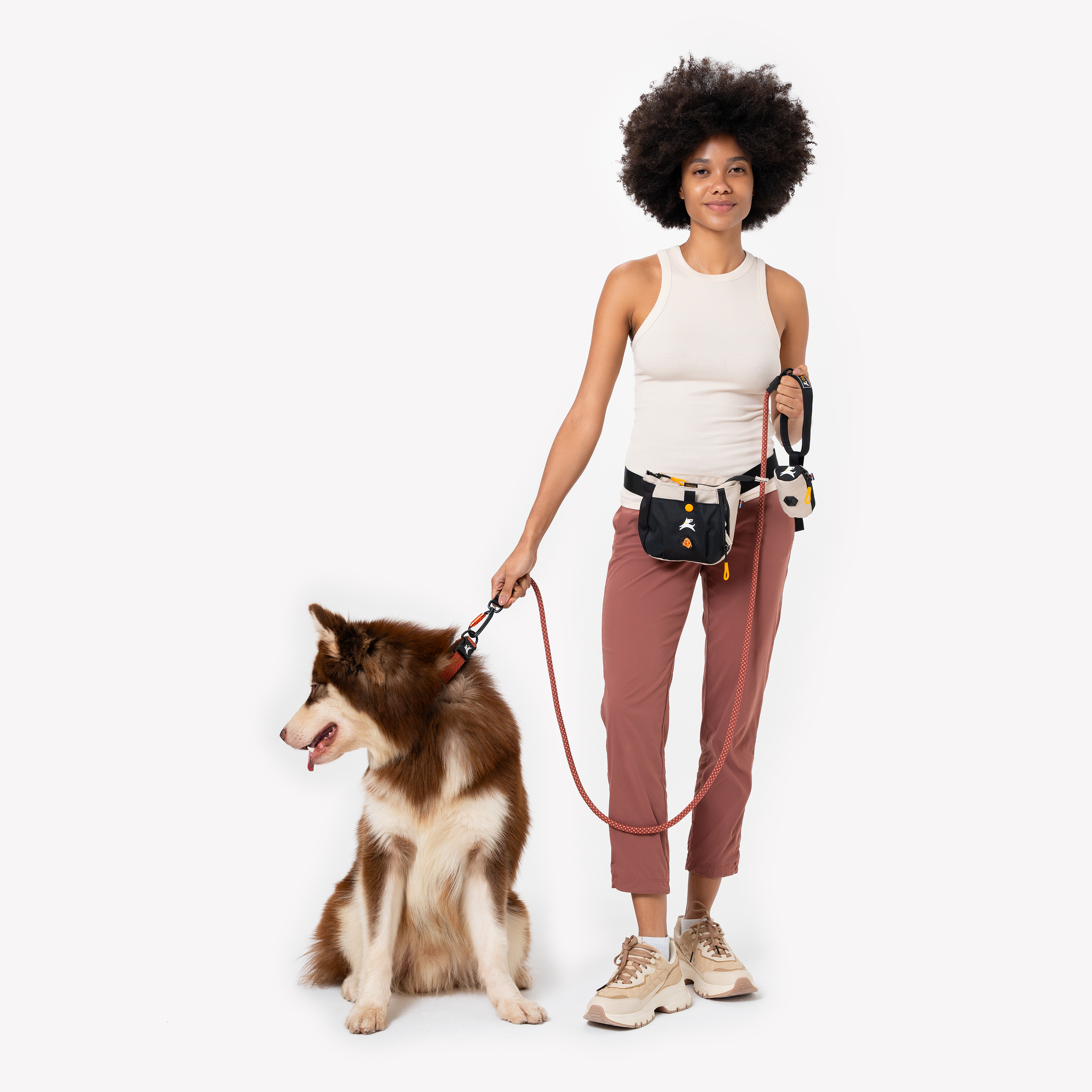 Woman walking a brown and white dog, wearing a beige and black waist bag with matching accessories, dressed in a cream tank top, mauve pants, and beige sneakers #color_con leche