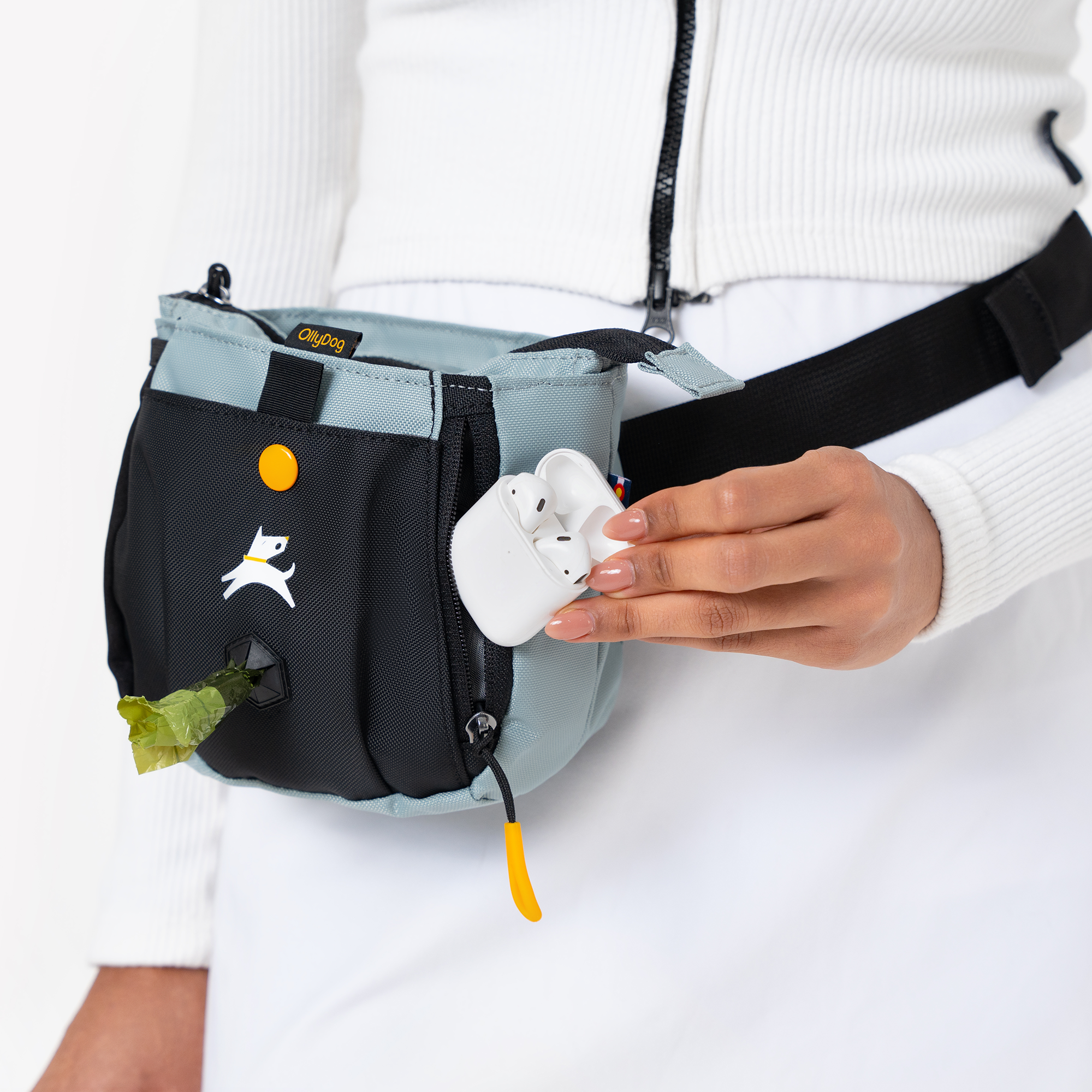 Close-up of a woman wearing a two-tone blue and black OllyDog waist bag, pulling a green waste bag from the dispenser and holding white wireless earbuds, dressed in a white zip-up top and skirt #color_st. lucia