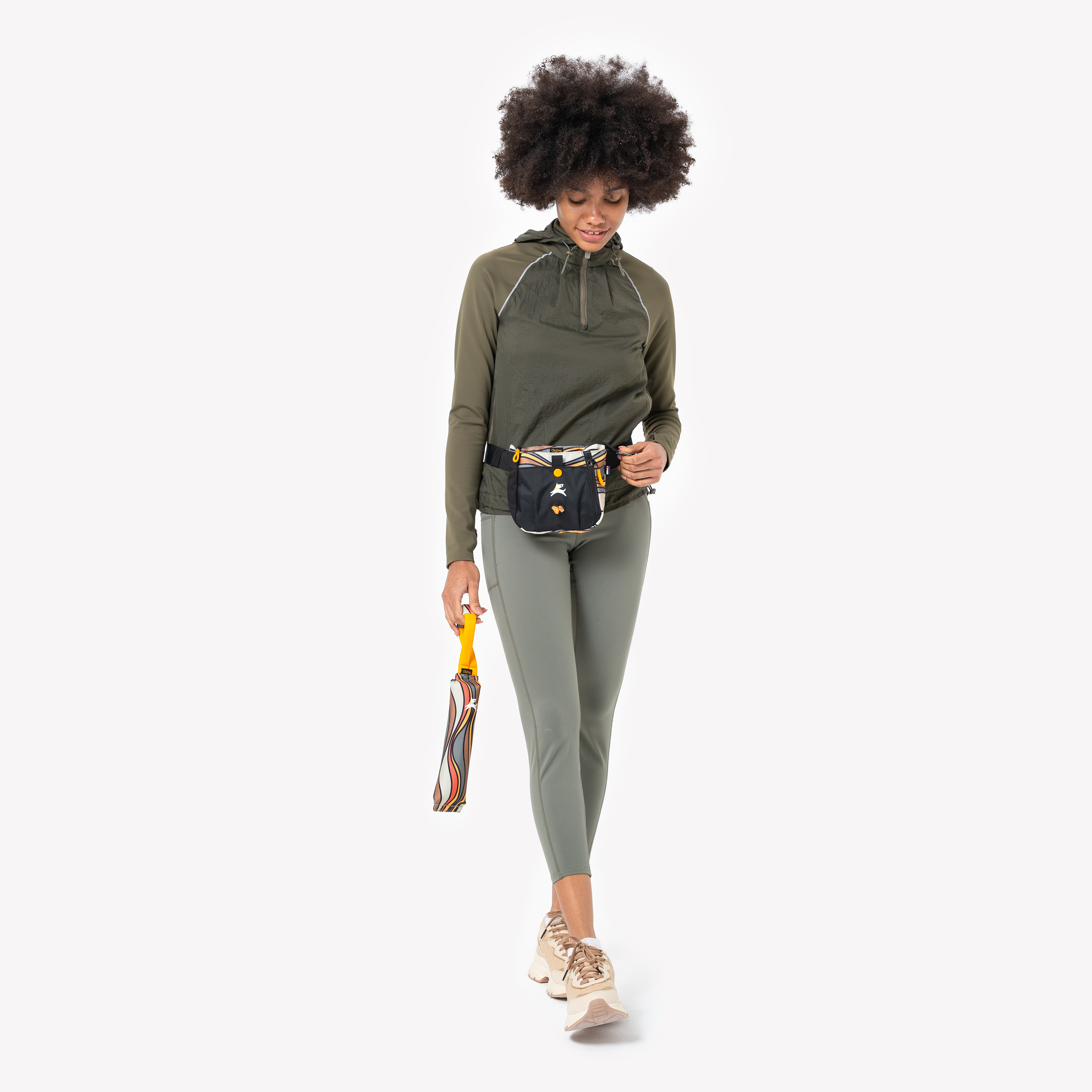 Person dressed in an olive green hoodie and leggings, wearing a black and orange dog walking waist bag and holding a colorful rolled-up leash, walking forward confidently #color_ventura '73
