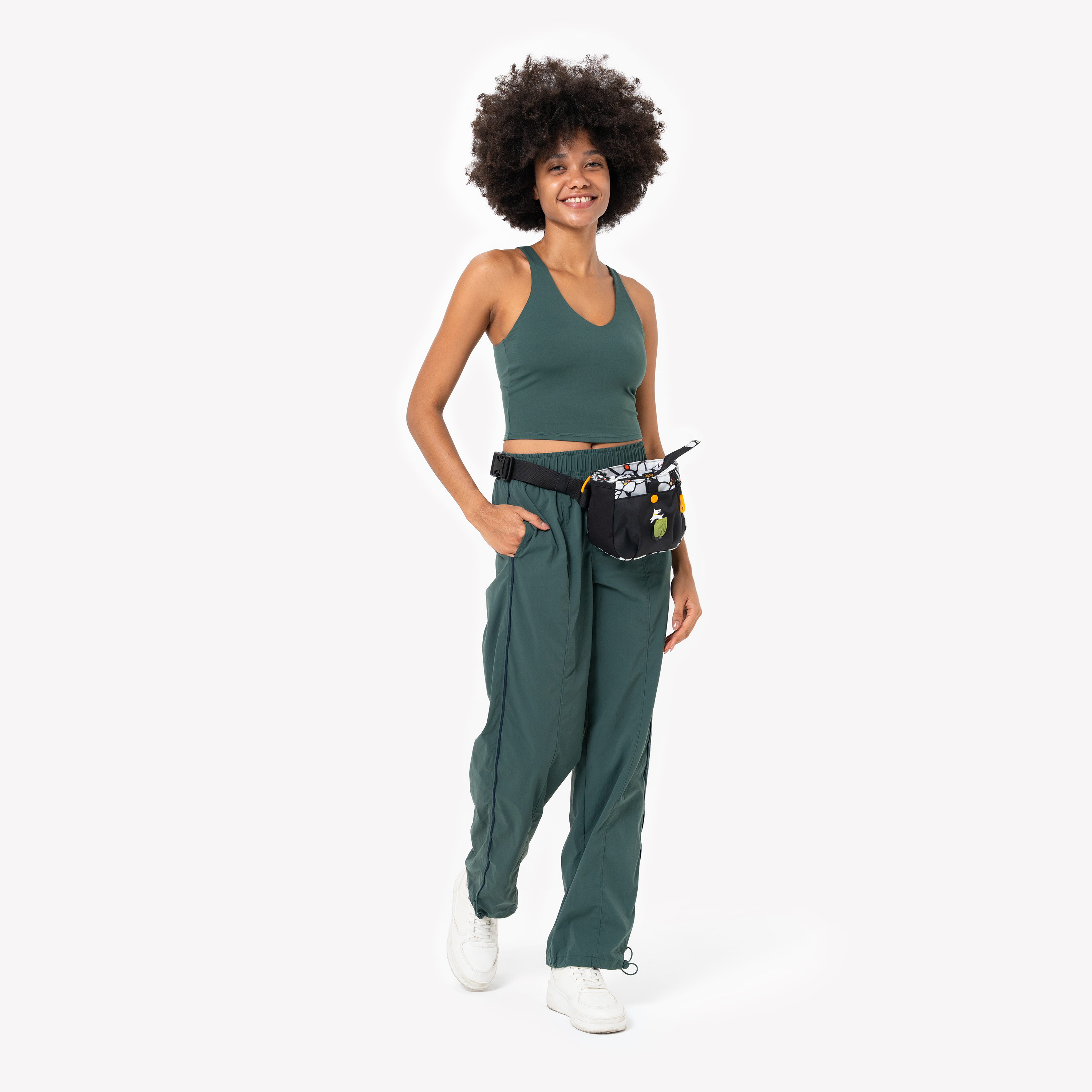 Woman wearing a green crop top and pants, smiling and posing with a black and patterned waist pouch designed for dog walking #color_magnolia